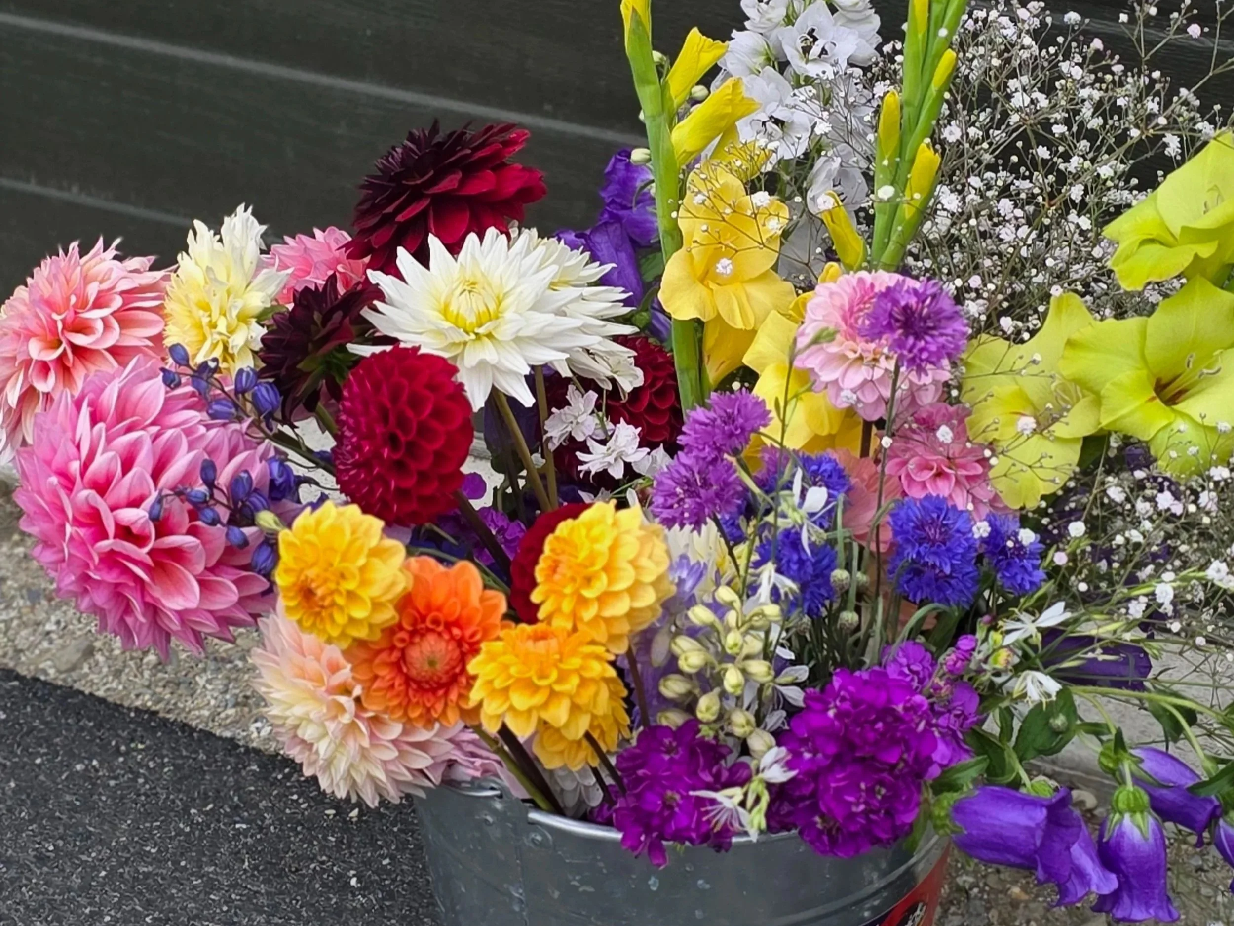 A large bouquet of colorful flowers, including dahlias, lilies, and other blossoms, arranged in a metal container on a rubber mat surface.