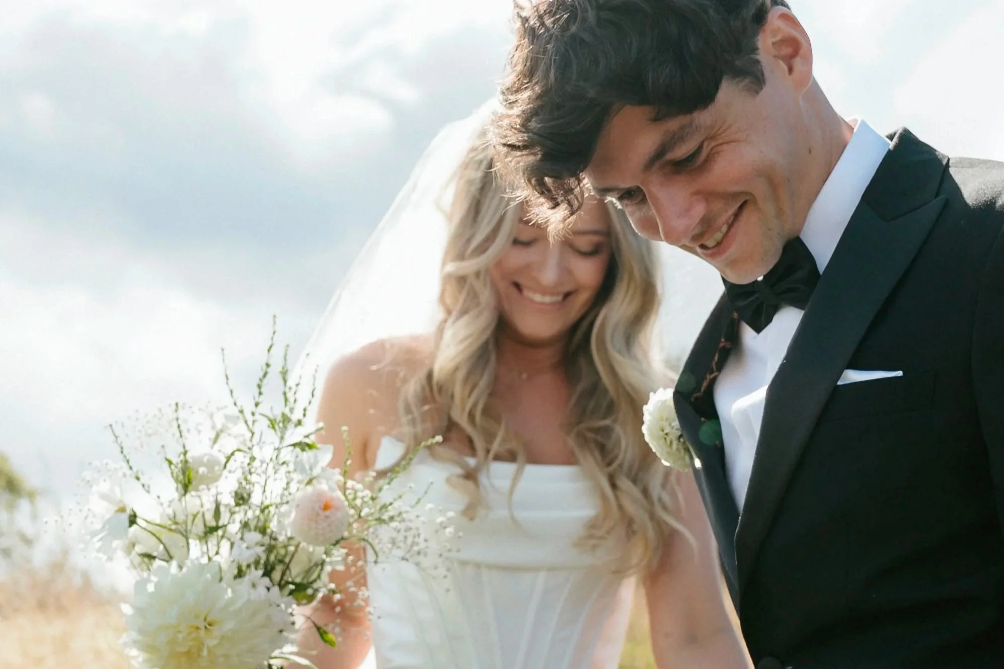 A bride and groom smiling and looking down, holding a bouquet of white flowers, outdoors on a cloudy day.
