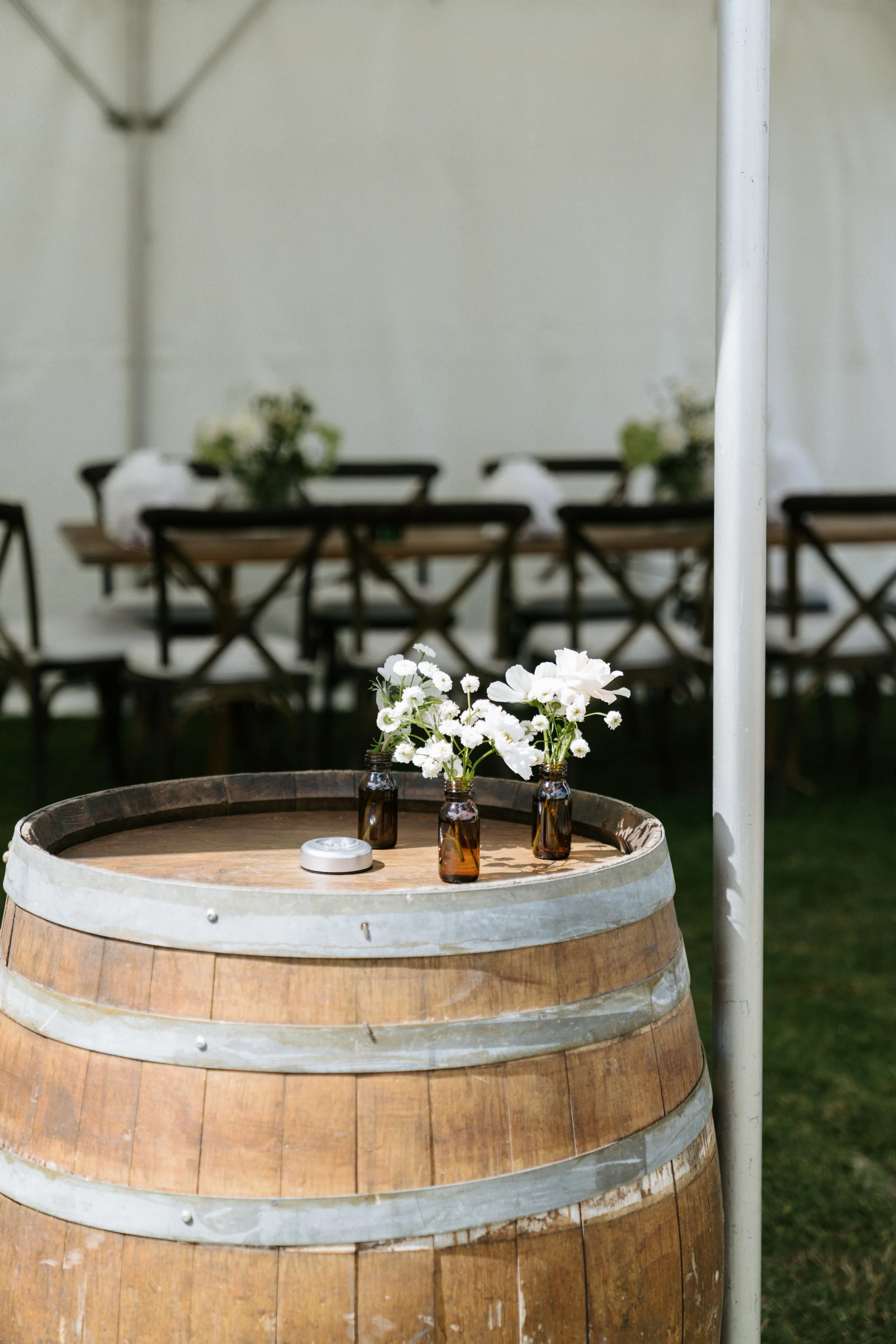 A round wooden barrel with silver metal bands, serving as a table, topped with three small brown glass bottles holding white daisy and flower arrangements, and a round white object, under a white shade umbrella, in an outdoor setting with tables, chairs, and floral decorations in the background.