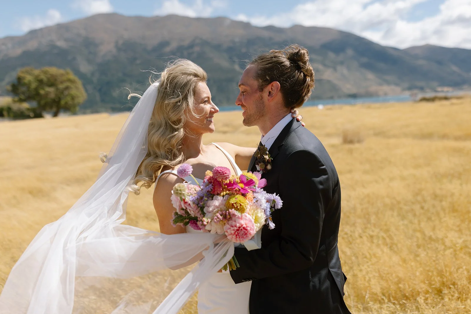 A bride and groom on their wedding day in a field with mountains in the background, smiling and looking at each other, with the bride holding a bouquet of pink, purple, and yellow flowers.