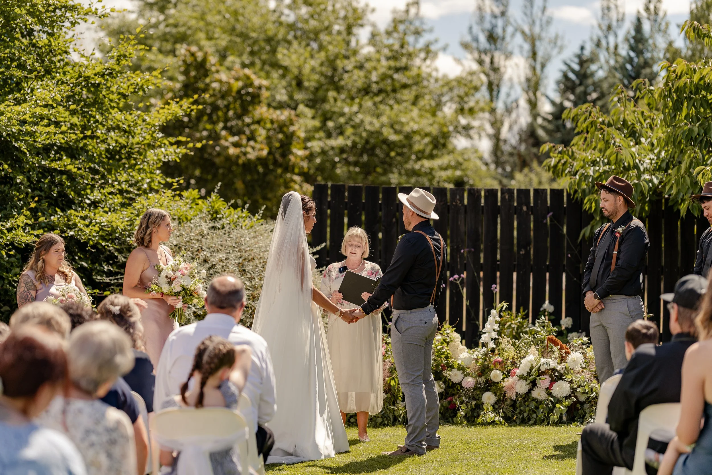 A wedding ceremony outdoors with the bride and groom holding hands, surrounded by guests and floral arrangements, and a officiant officiating.