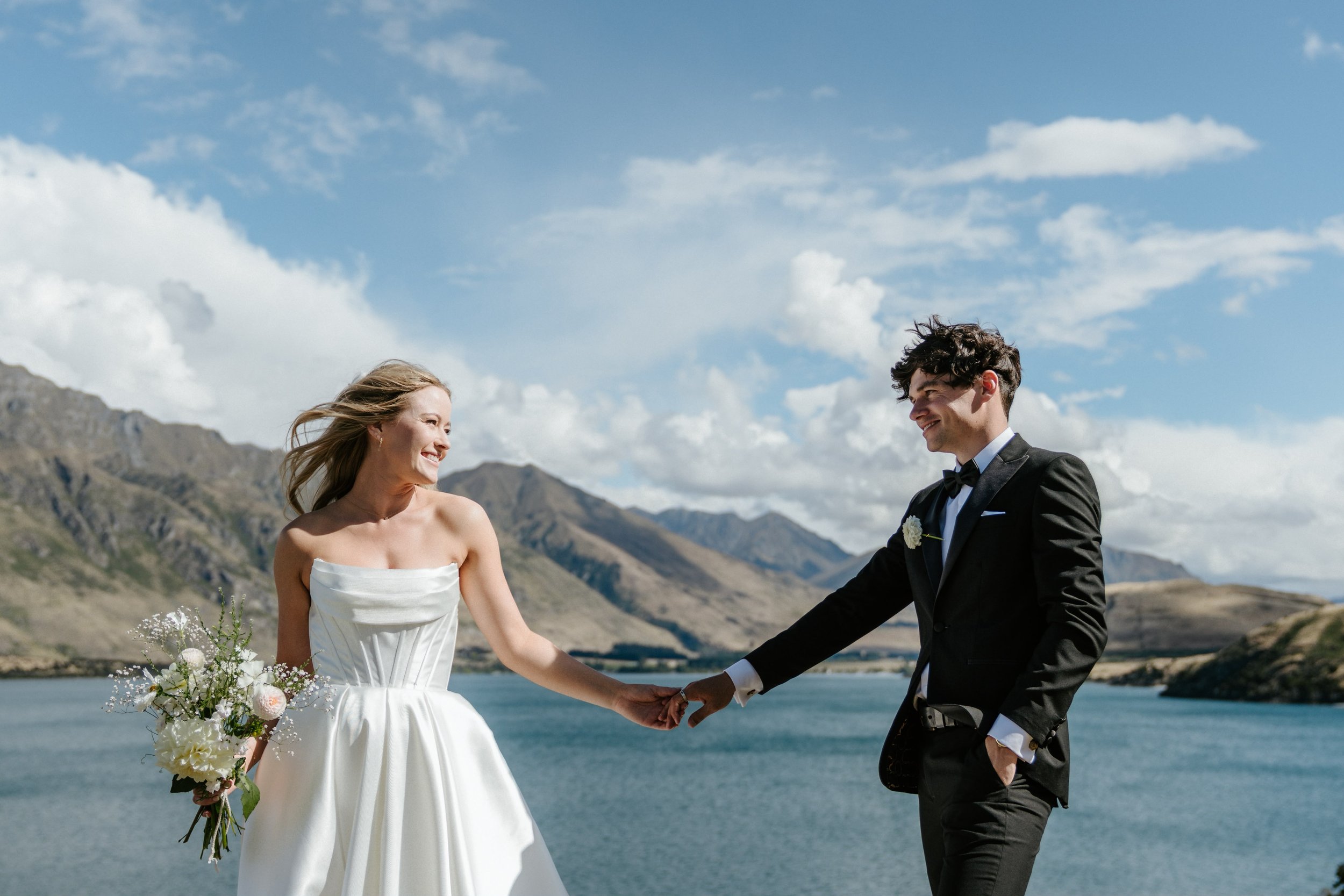 A bride and groom holding hands and smiling at each other outdoors by a lake, with mountains and cloudy sky in the background.