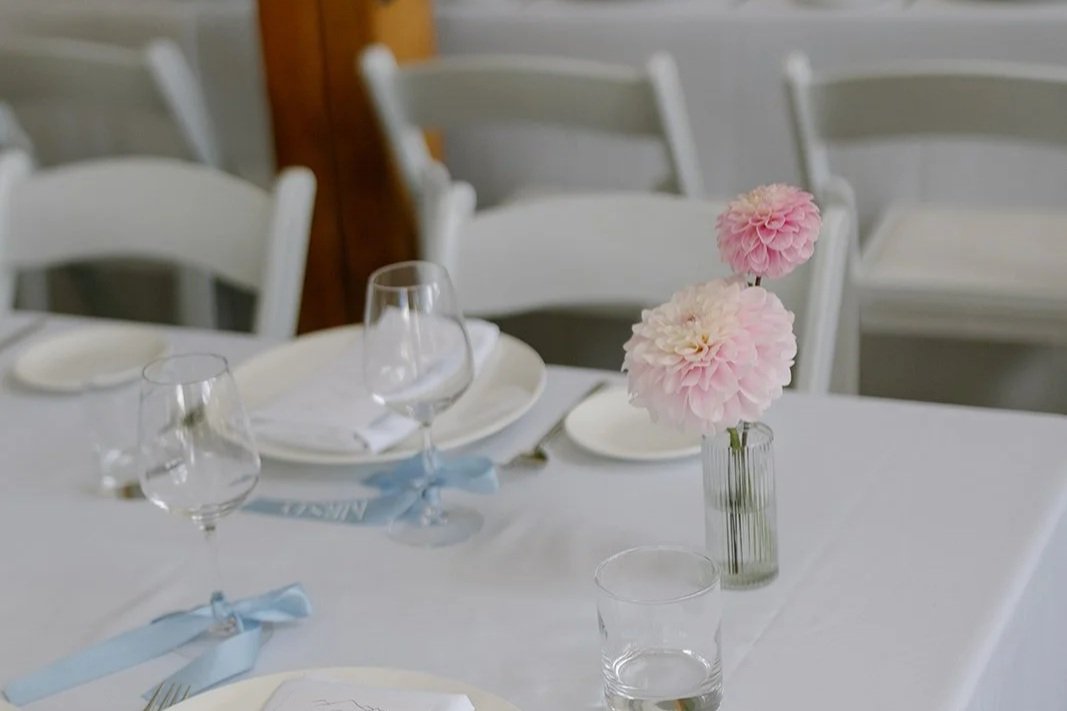 A table set for a formal event with pink dahlia flowers in a transparent vase, white plates, and wine glasses, with white chairs in the background.