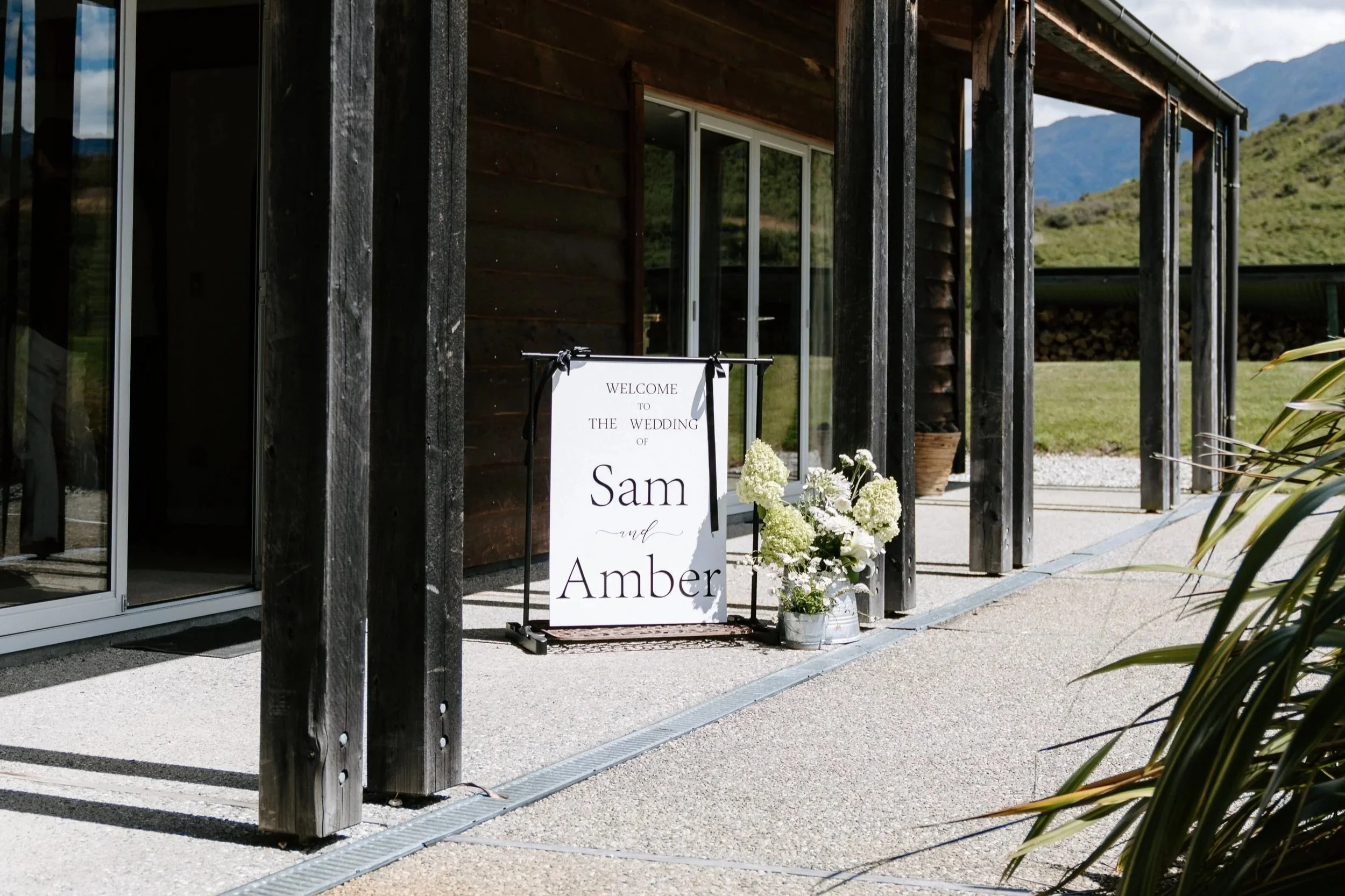 Wedding welcome sign with flowers at a rustic building in an outdoor setting with mountains in the background.