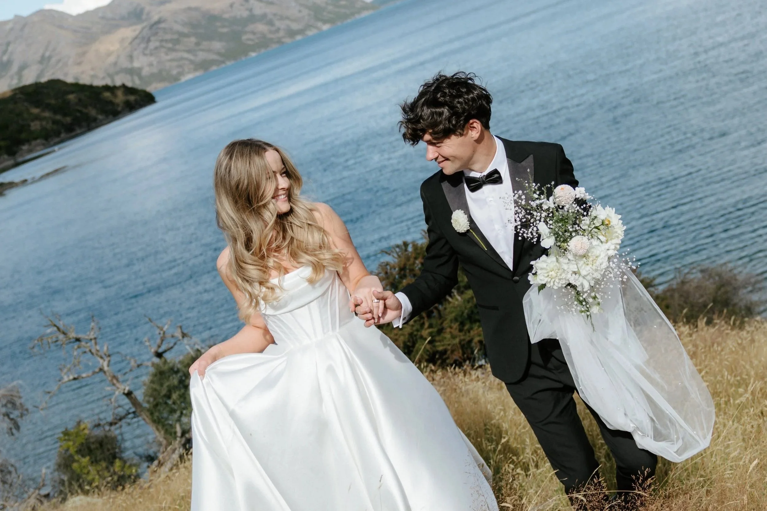 A bride and groom holding hands and smiling by a body of water with mountains in the background, during their wedding.