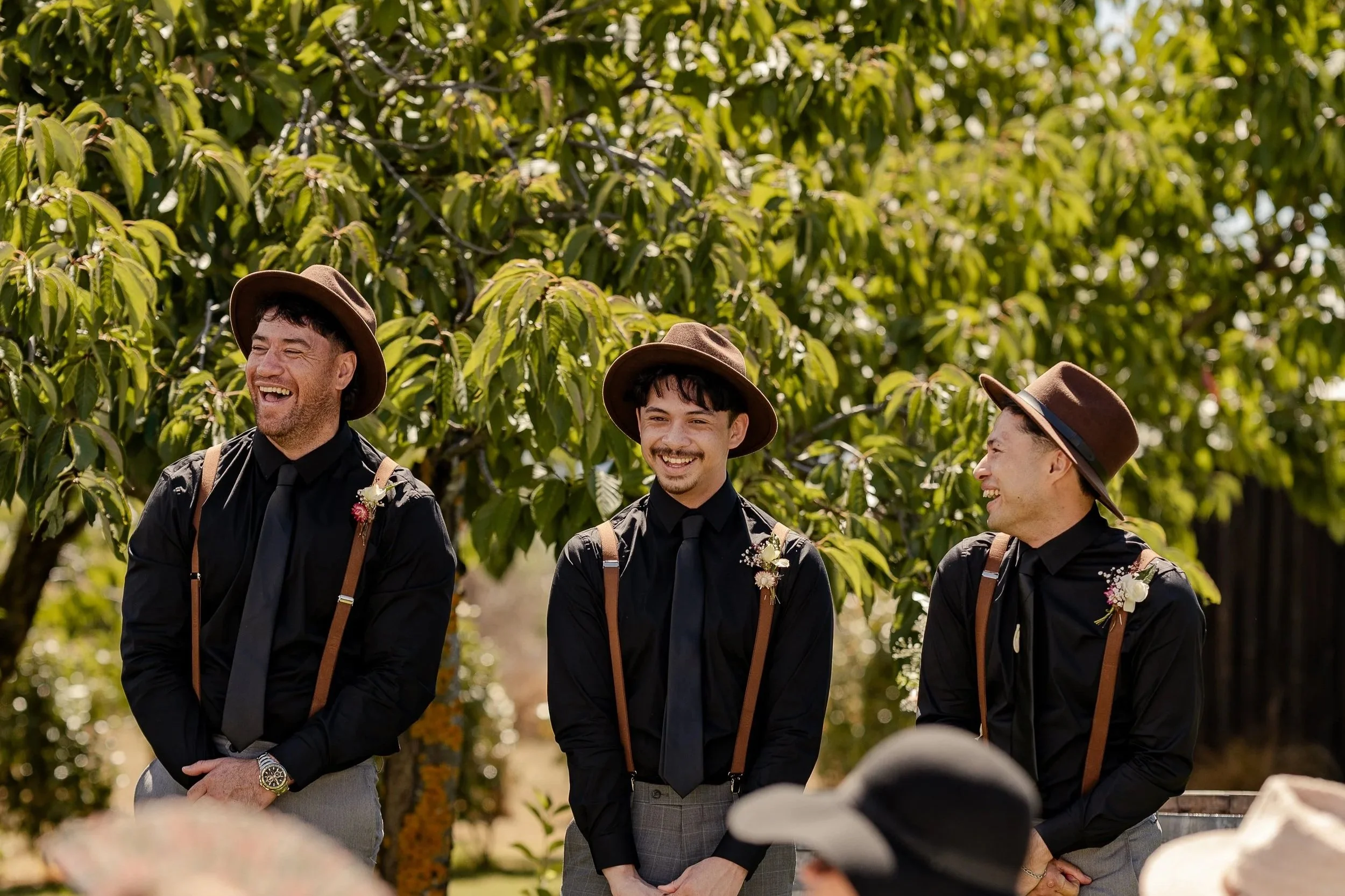 Three men dressed in black shirts and brown hats, smiling and standing outdoors amid green foliage, possibly at a garden or park.