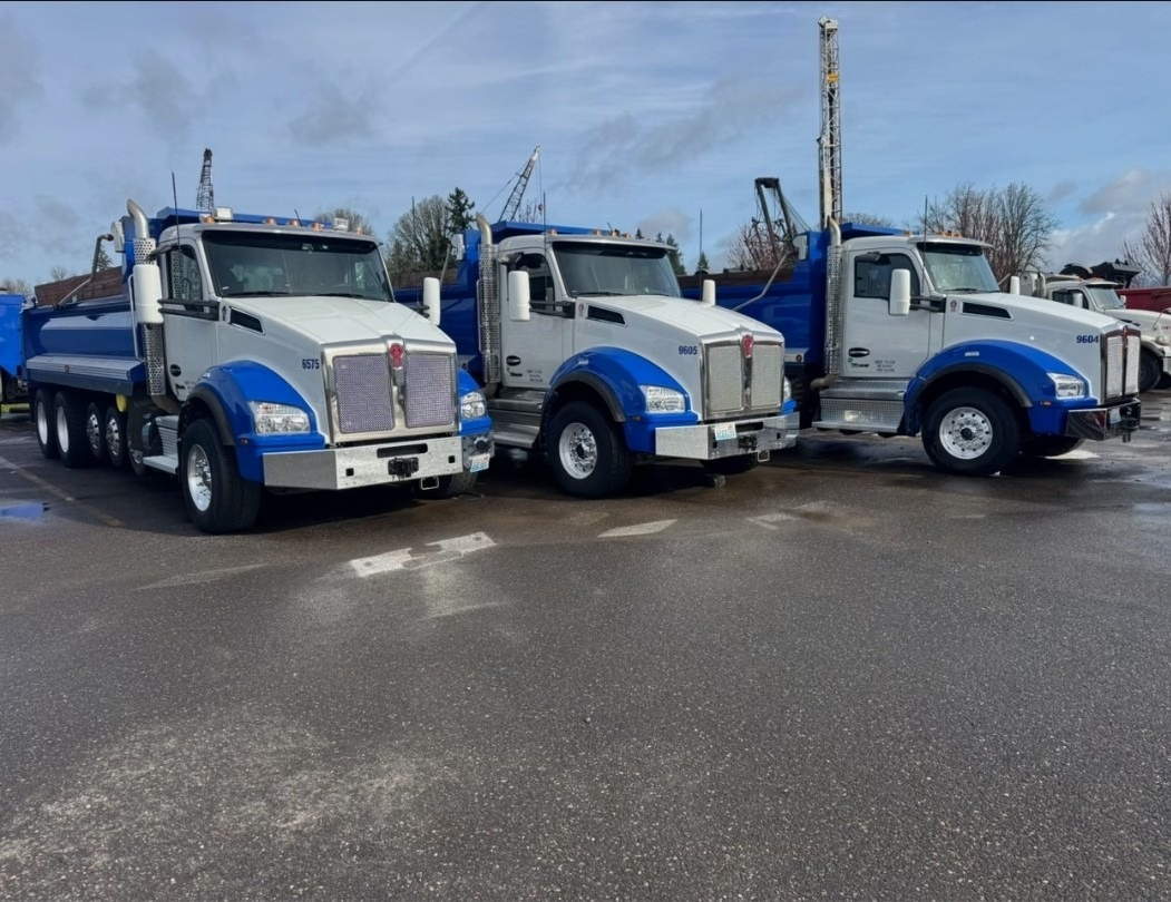Three large work trucks parked on a wet pavement, with cranes visible in the background.