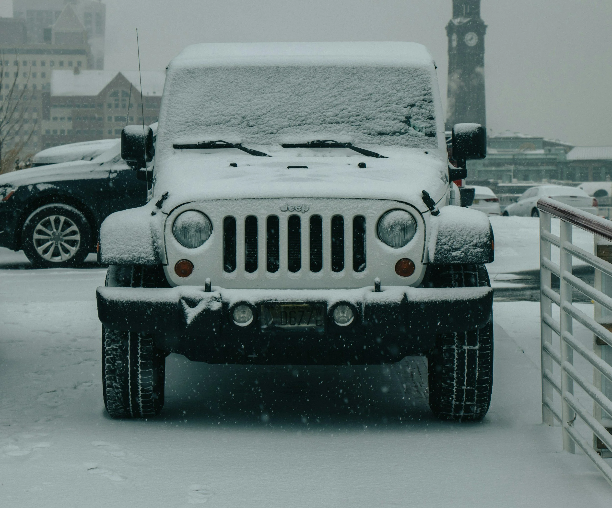 Front view of a Jeep vehicle covered in snow, parked outdoors in a snowy city scene with buildings in the background.