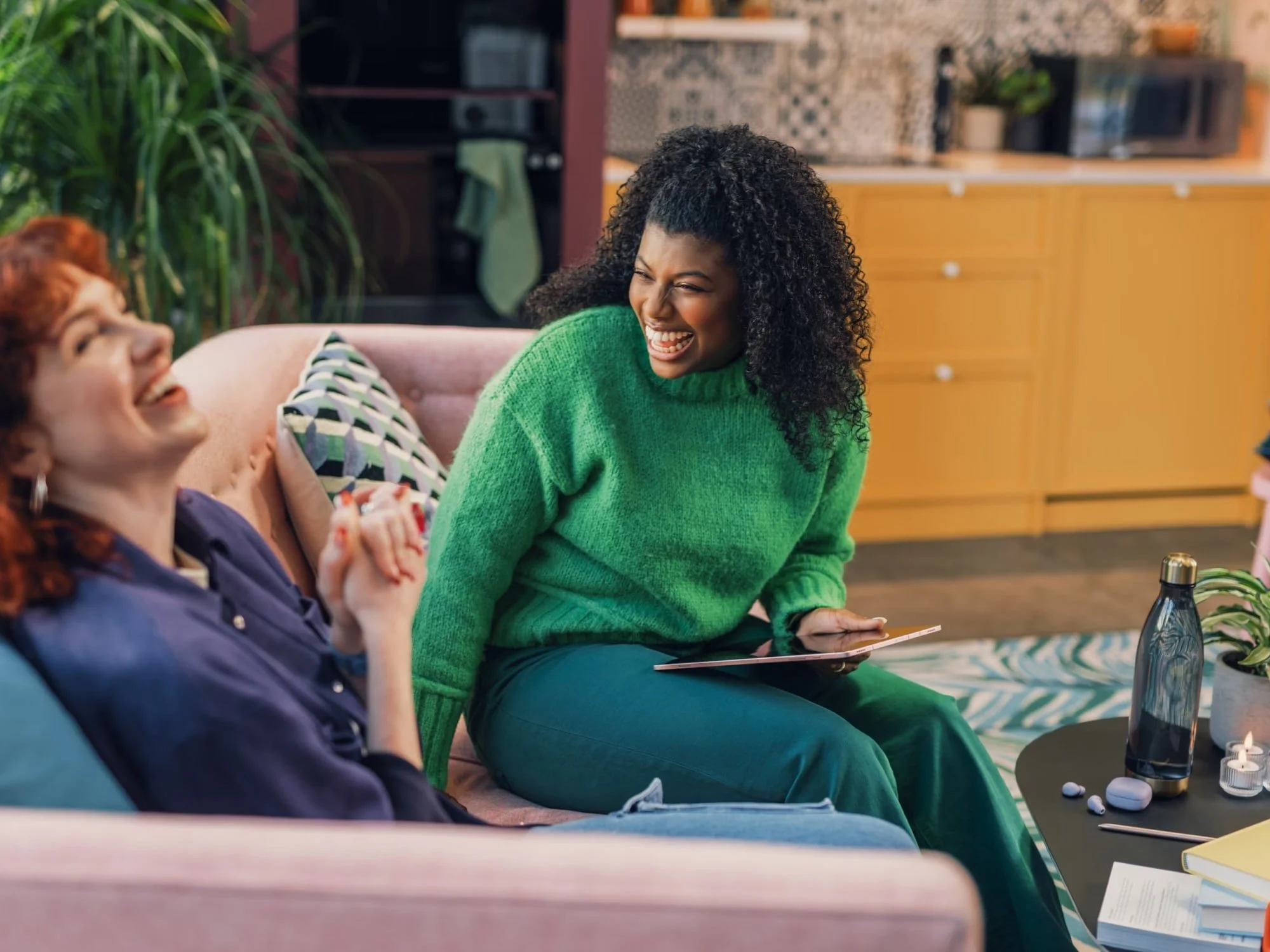 Two women sitting on a couch, laughing and holding hands indoors, with a coffee table nearby.