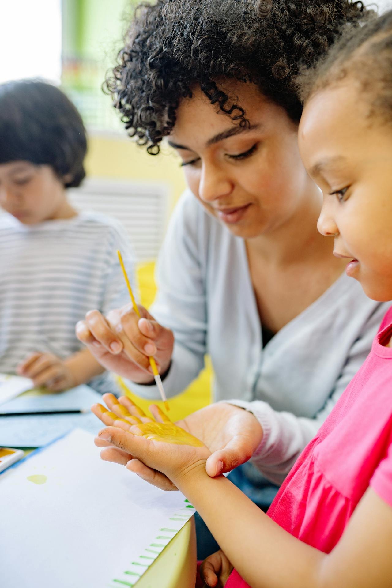 A woman and a young girl are engaged in a painting activity at a table. The woman is applying yellow paint to the girl's hand with a small brush, while the girl is holding her hand out, observing the process. In the background, another child is seen, slightly out of focus.
