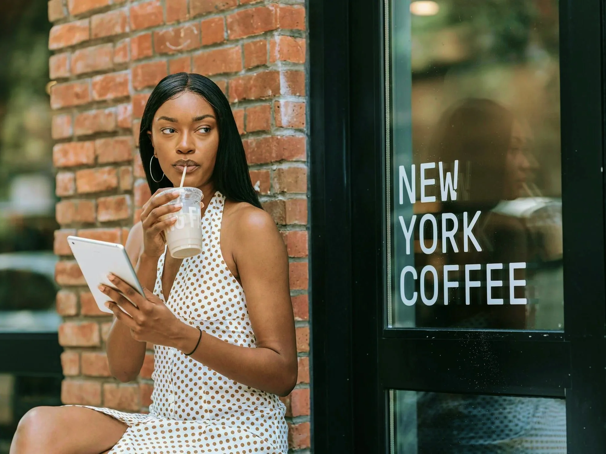 A young woman in a polka dot dress drinks a coffee beverage and looks at her phone outside a café with a large window that has 'New York Coffee' written on it.
