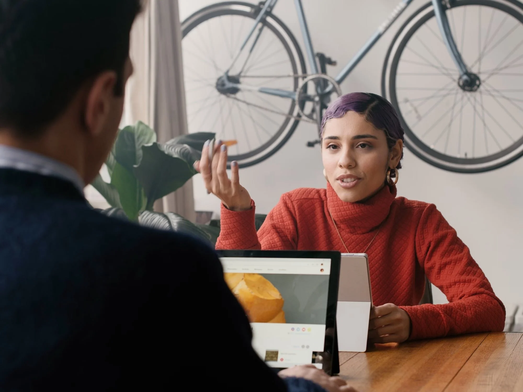 A woman with short purple hair and large gold earrings is speaking during a conversation with a man in a dark blazer. They are seated at a wooden table with laptops in front of them. A bicycle is mounted on the wall in the background, and there is a large leafy plant nearby.