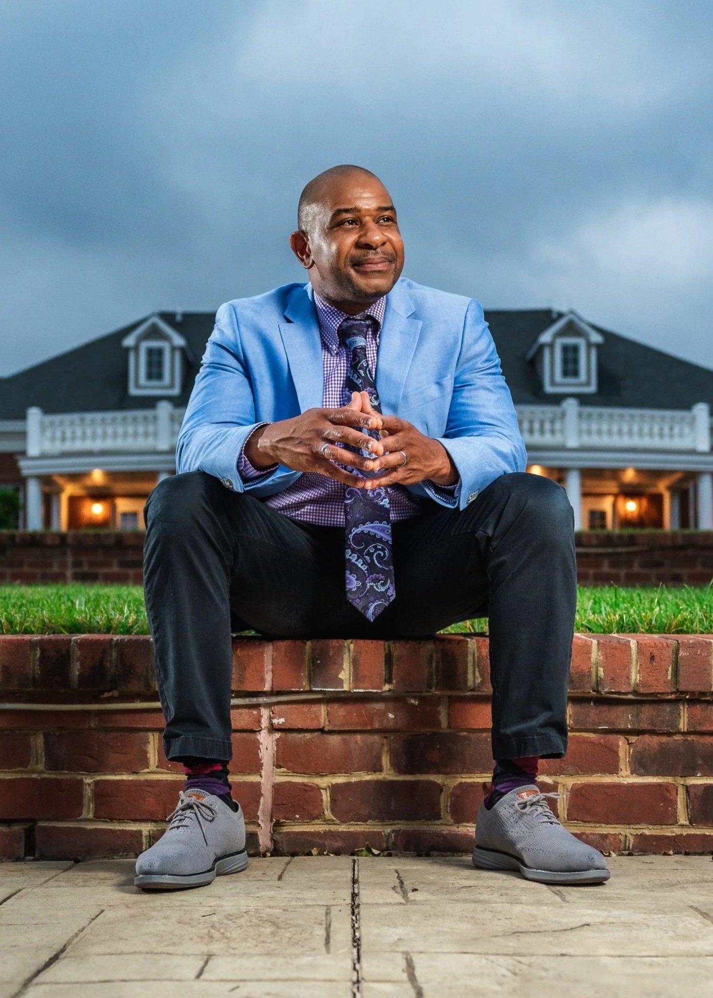 A man in a light blue blazer, dress shirt, and patterned tie sitting on a brick ledge outside a large house with a porch, under cloudy sky.