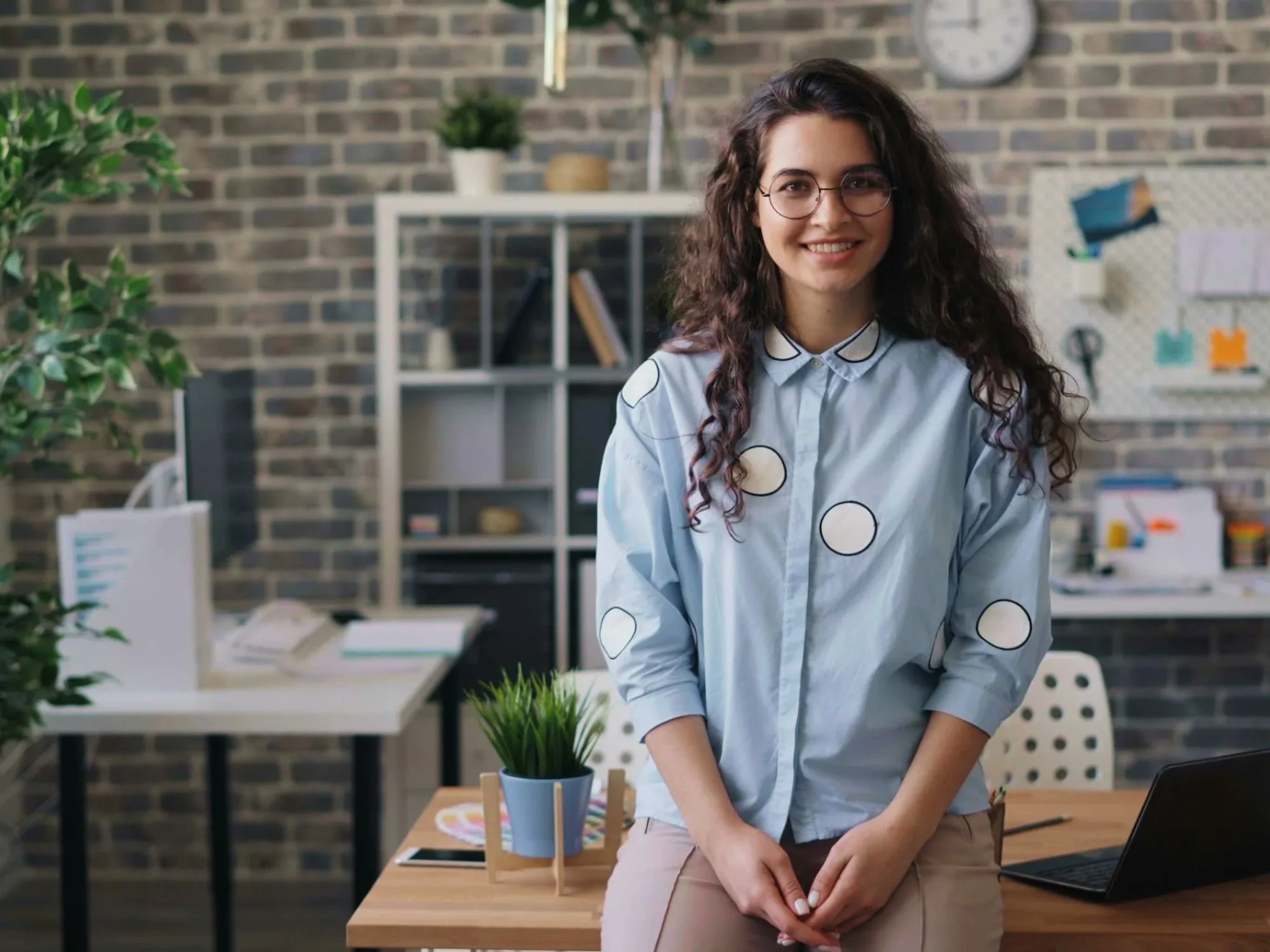 A smiling woman with curly hair and glasses standing in an office with a brick wall background, plants, a white shelf, a desk with a computer, and a notebook.