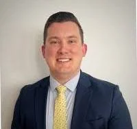 Close-up of a smiling woman with curly hair, wearing glasses, earrings, a necklace, and a striped shirt, in a bright room.