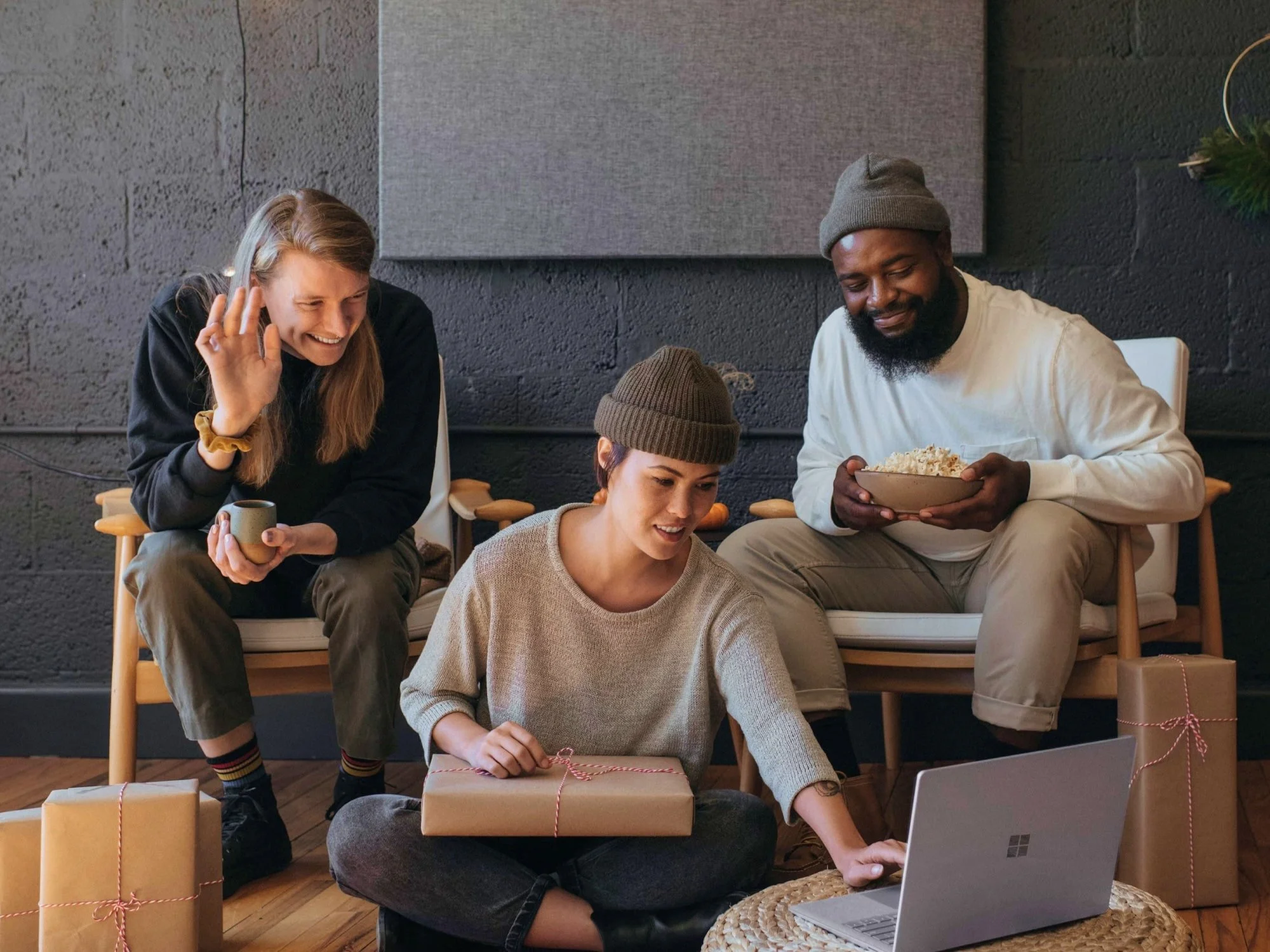 Three friends celebrating, opening gifts, and watching something on a laptop in a cozy room with wrapped presents around.