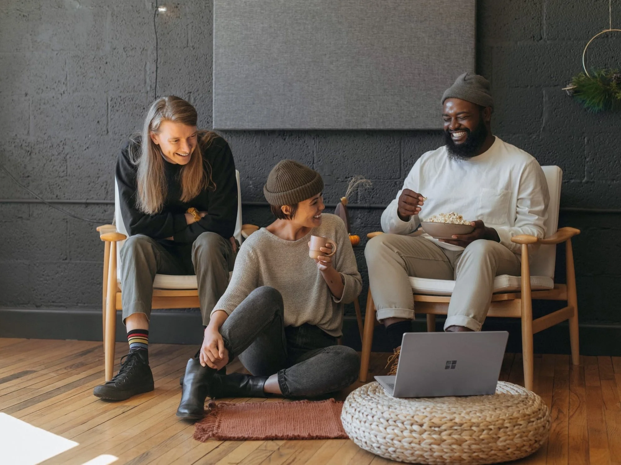 Three people sitting in a cozy room with a dark gray wall, enjoying snacks and drinks while watching something on a laptop placed on a round woven ottoman. One person is sitting on a chair, another is seated on the floor, and the third is in a chair.