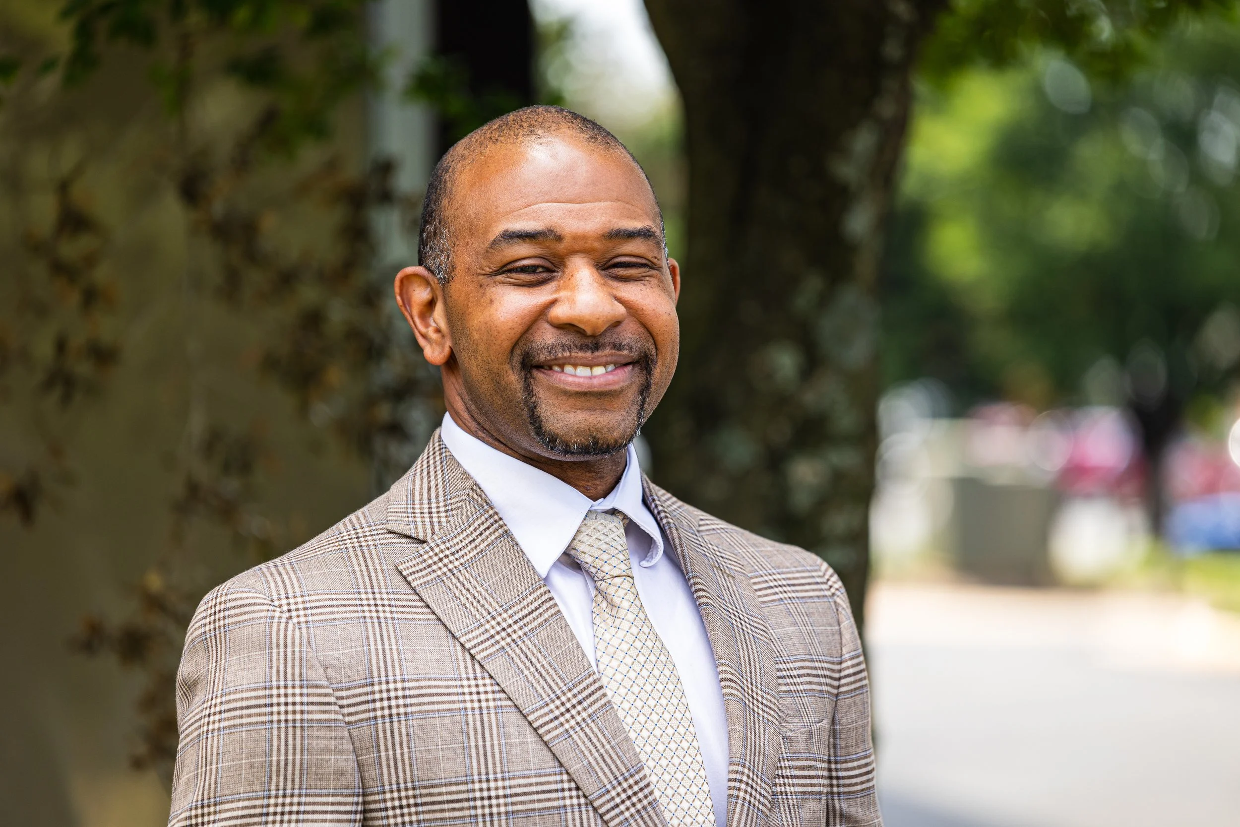 A smiling man in a plaid suit and cream tie standing outdoors with trees in the background.