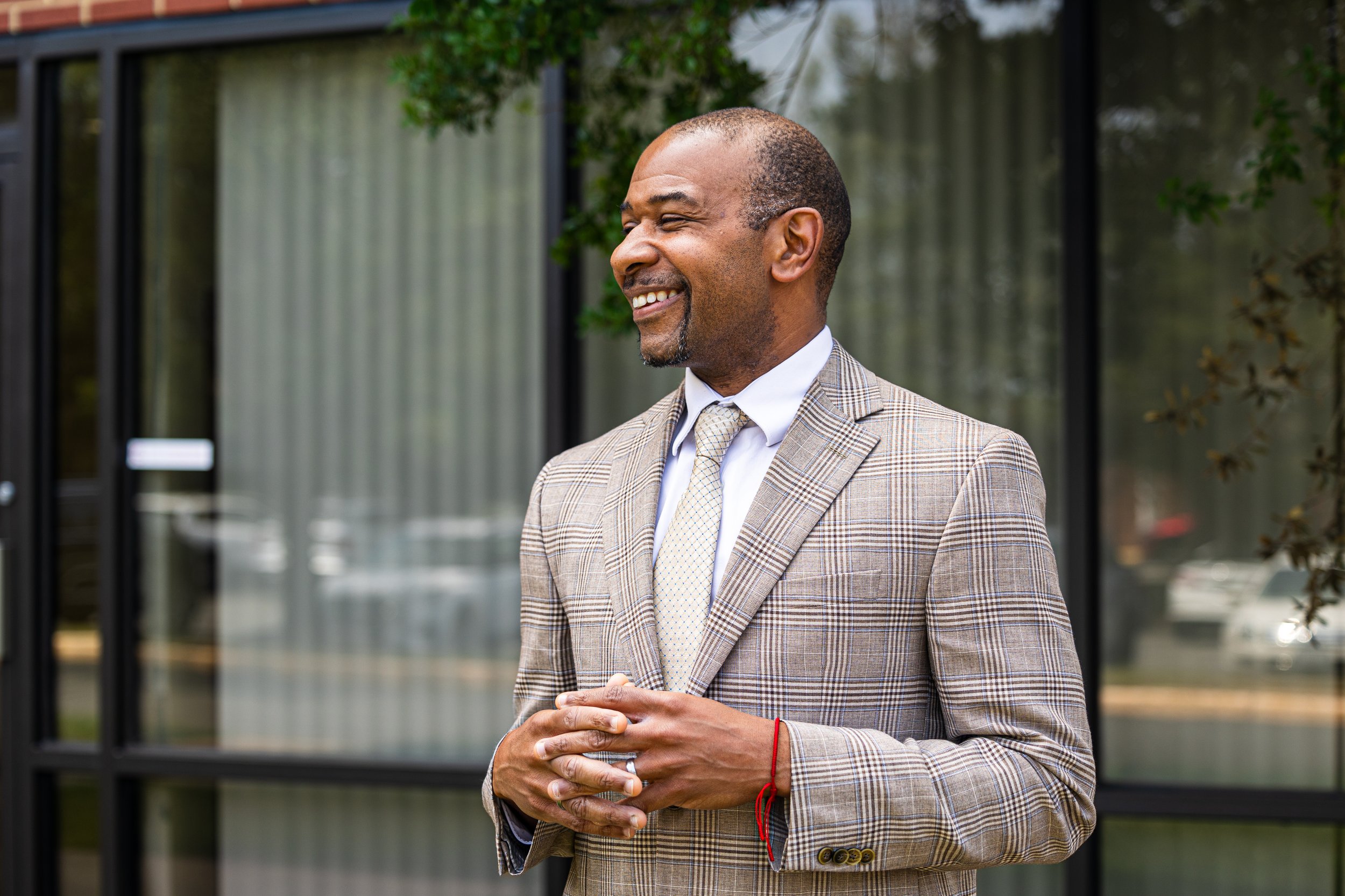 A smiling man in a brown plaid suit and tie standing outside in front of a glass building with trees.