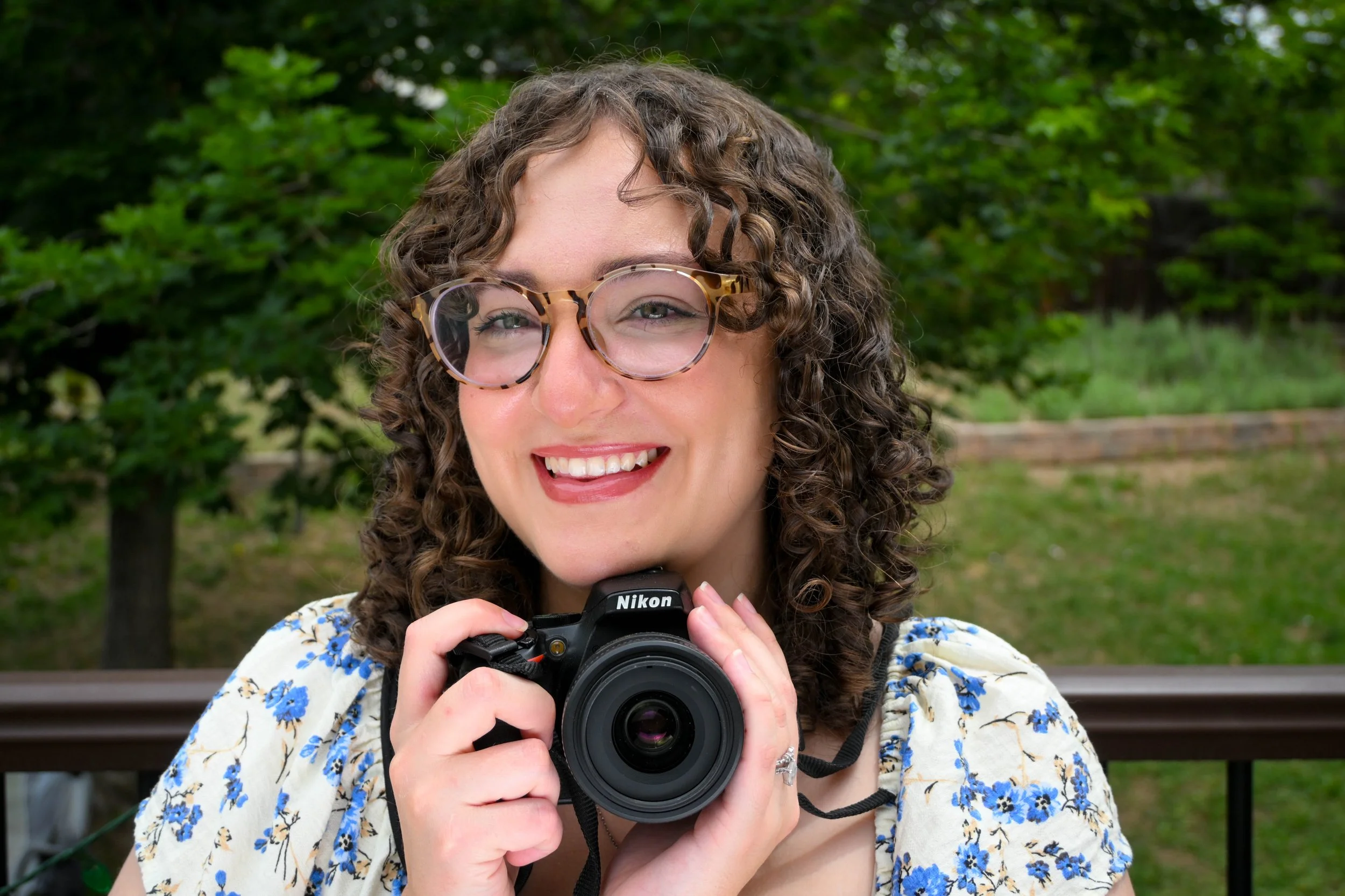 A woman photographer in Centennial, Colorado with curly hair and glasses smiling while holding a Nikon camera outdoors with green trees in the background.