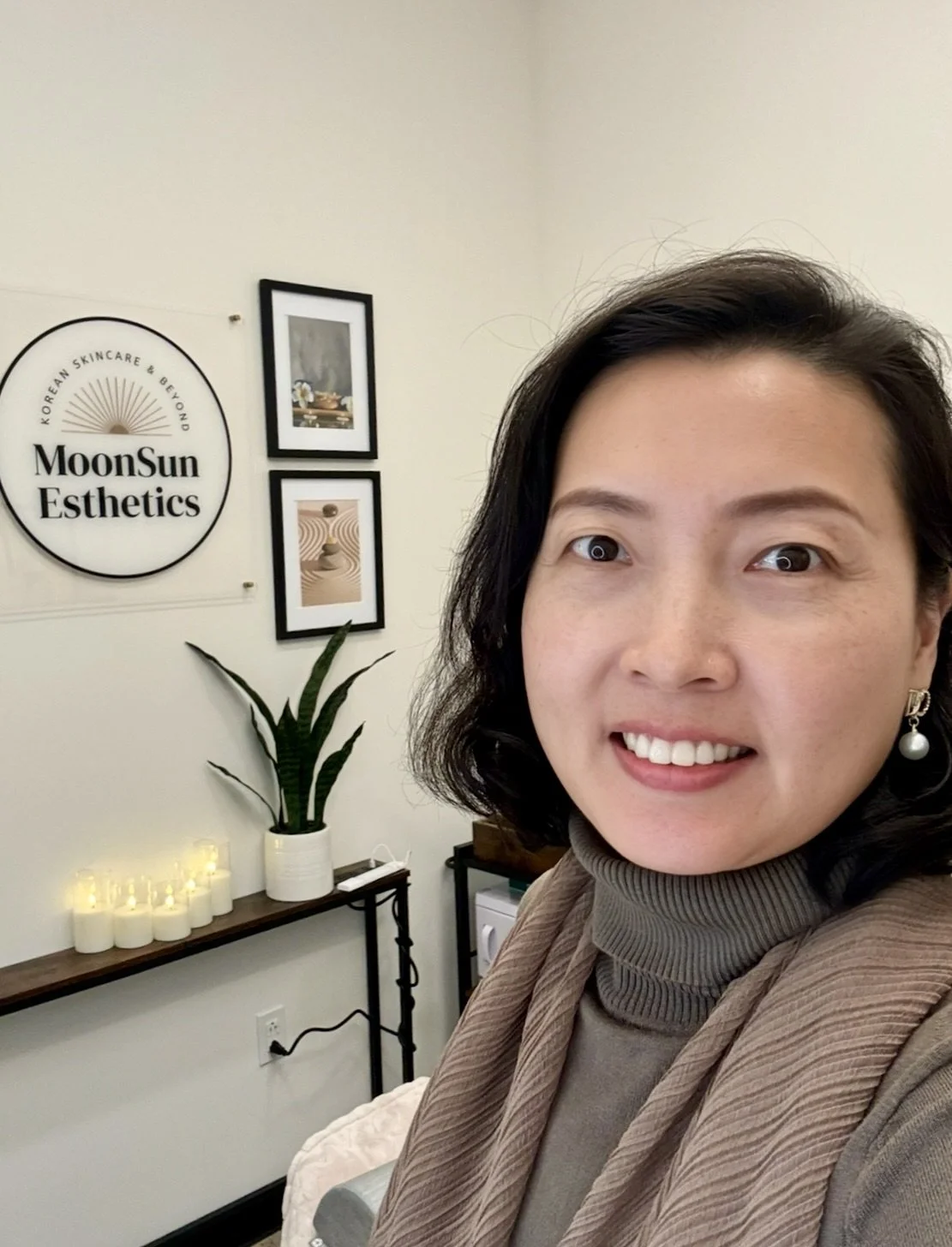 The founder of MoonSun Esthetics with black hair and earrings, smiling in a room with framed pictures, candles, a plant, and a sign that reads 'MoonSun Esthetics'.