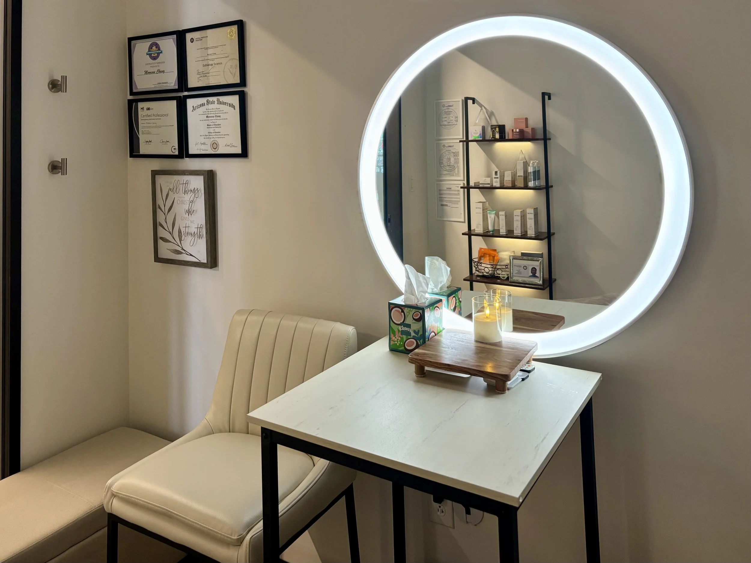 
At MoonSun Esthetics - A cozy room corner with a white leather bench, a white table holding tissue and a wooden tray, and a large round illuminated mirror on the wall. The wall features framed certificates and artwork.