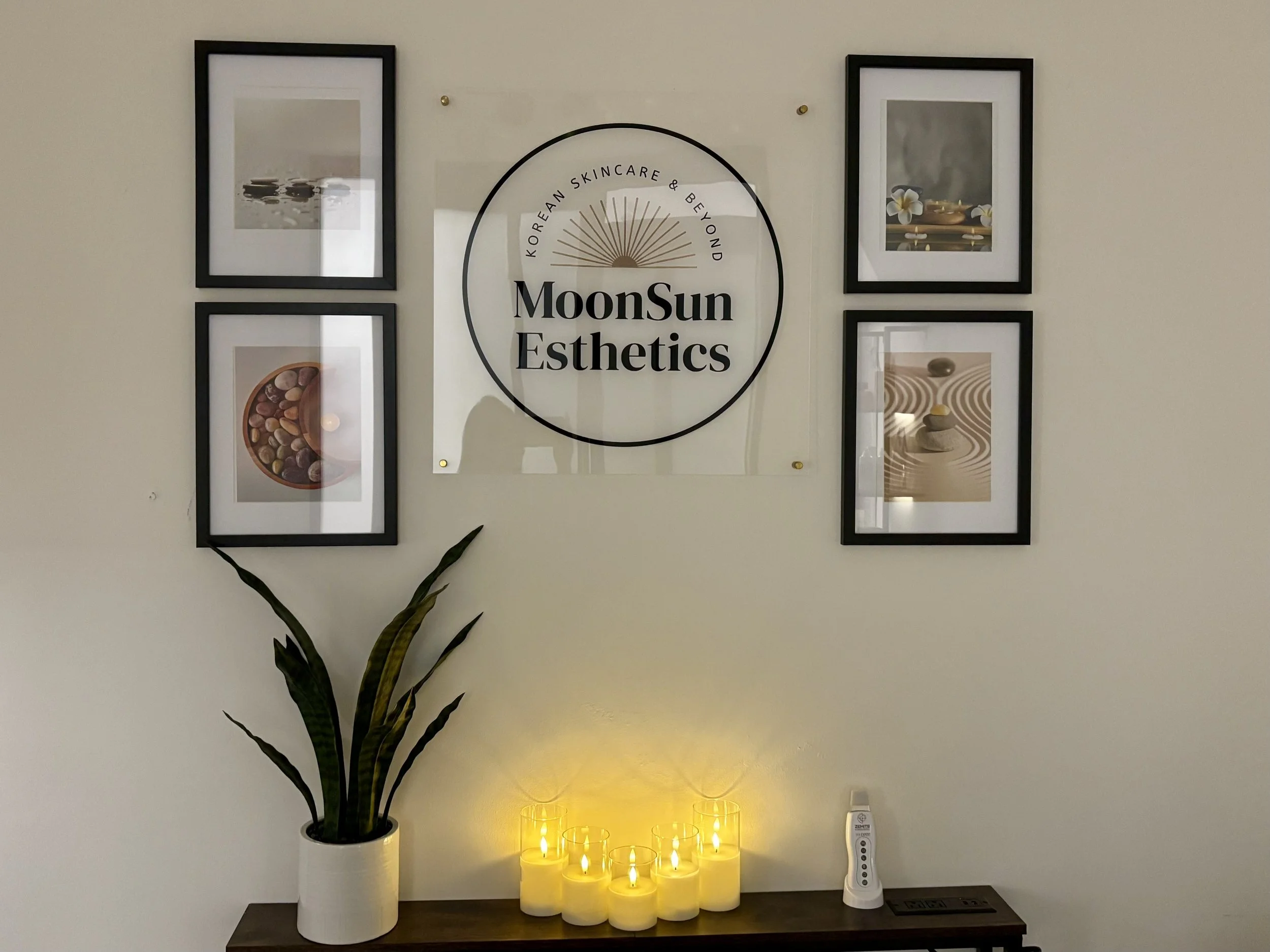 An interior wall decorated with four framed pictures and a circular sign reading 'Korean Skincare & Beyond MoonSun Esthetics.' Below the sign, a potted plant with long, green leaves stands on a table. Several yellow candle-like lights are illuminated