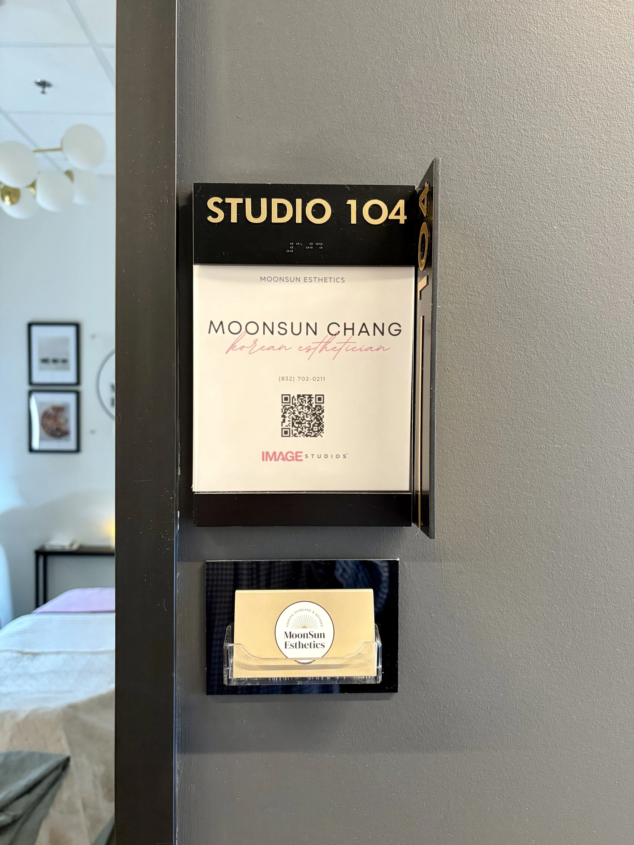 Sign on a gray wall that reads 'STUDIO 104' with Braille, below it a menu for Moonsun Esthetics with the name Moonsun Chang and their phone number, and a QR code. Below that, a business card for MoonSun Esthetics in a clear holder.