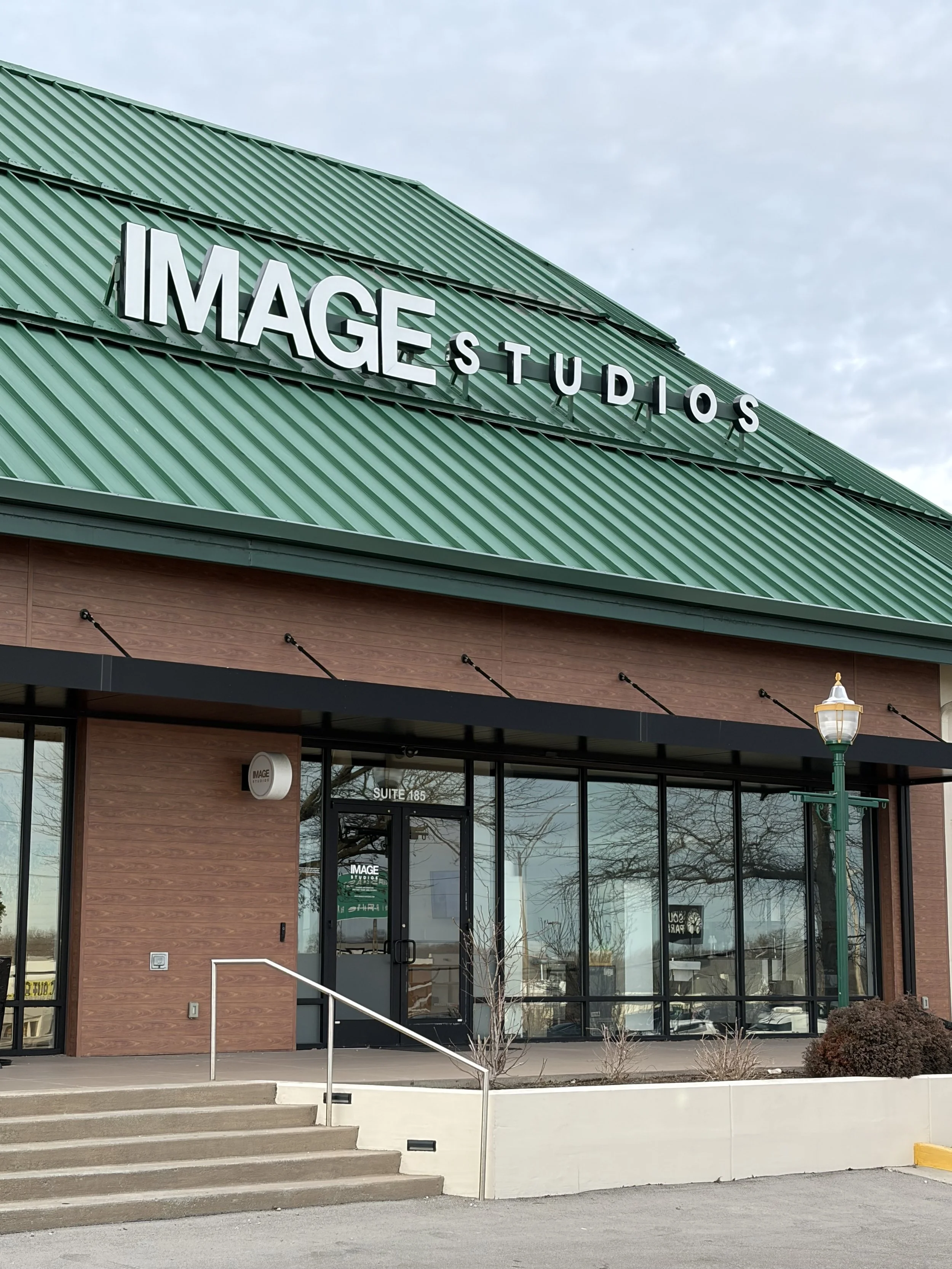 Exterior of a building with a green metal roof that has a large sign reading 'IMAGE STUDIOS' on it. The building has glass doors and windows, a brick facade, and a black awning. There is a small lamppost in front and a few small bushes near the sidew