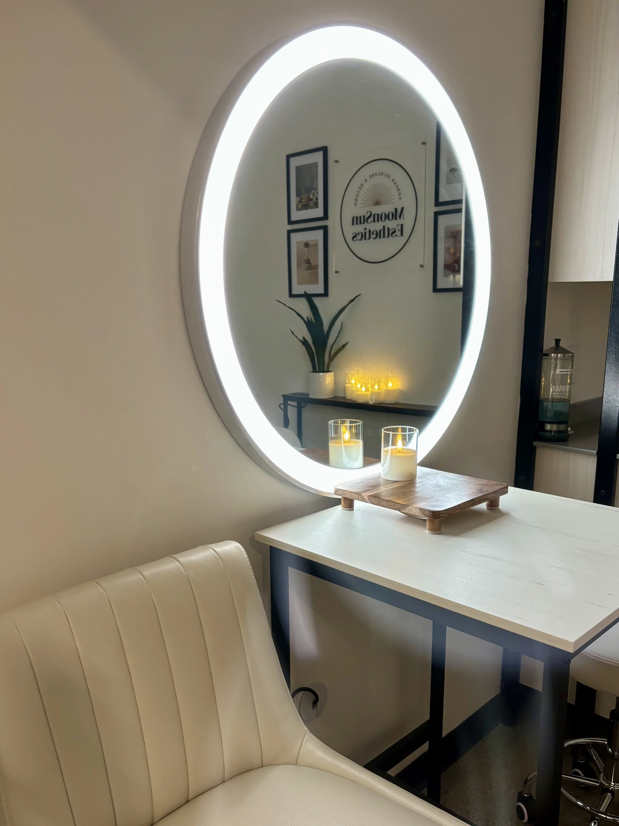 A large illuminated oval mirror reflecting a wall with framed pictures, a sign reading 'Moonsus Esthetics,' and a table with LED candles and a potted plant. There is a white cushioned chair in the foreground and a desk with a container labeled 'Barb'