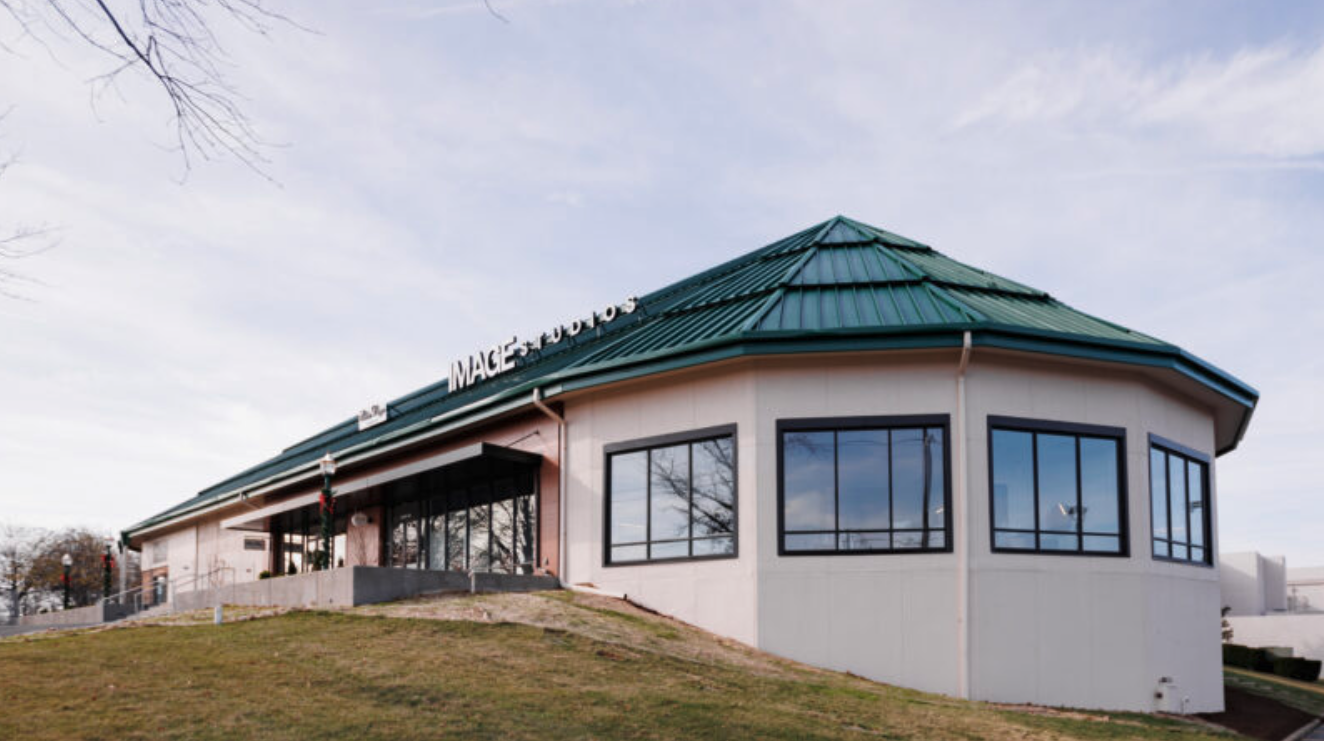At MoonSun Esthetics - A building with a green metal roof and large black-framed windows, situated on a grassy hill with a few leafless trees nearby and a cloudy sky in the background.