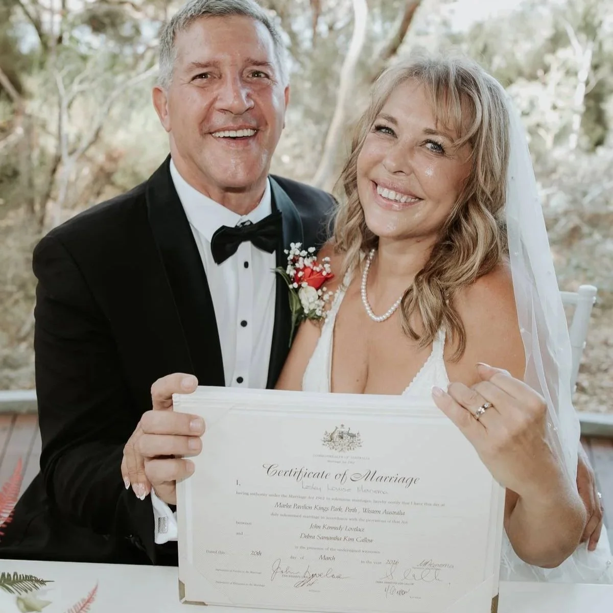 A happy bride and groom holding a marriage certificate outdoors, smiling and dressed in wedding attire.