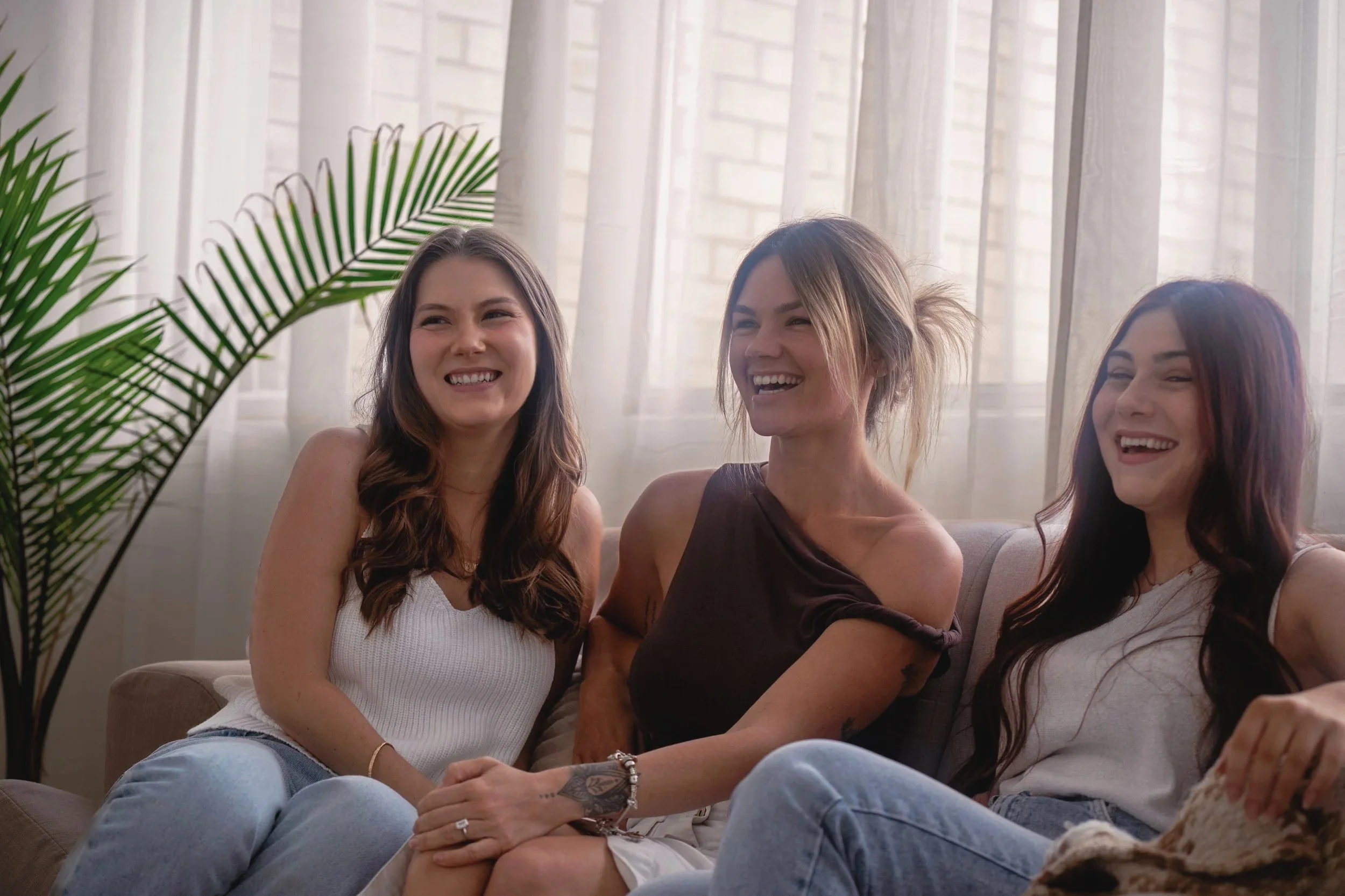Three women sitting on a beige couch, smiling and laughing, with sheer white curtains and a potted plant in the background.