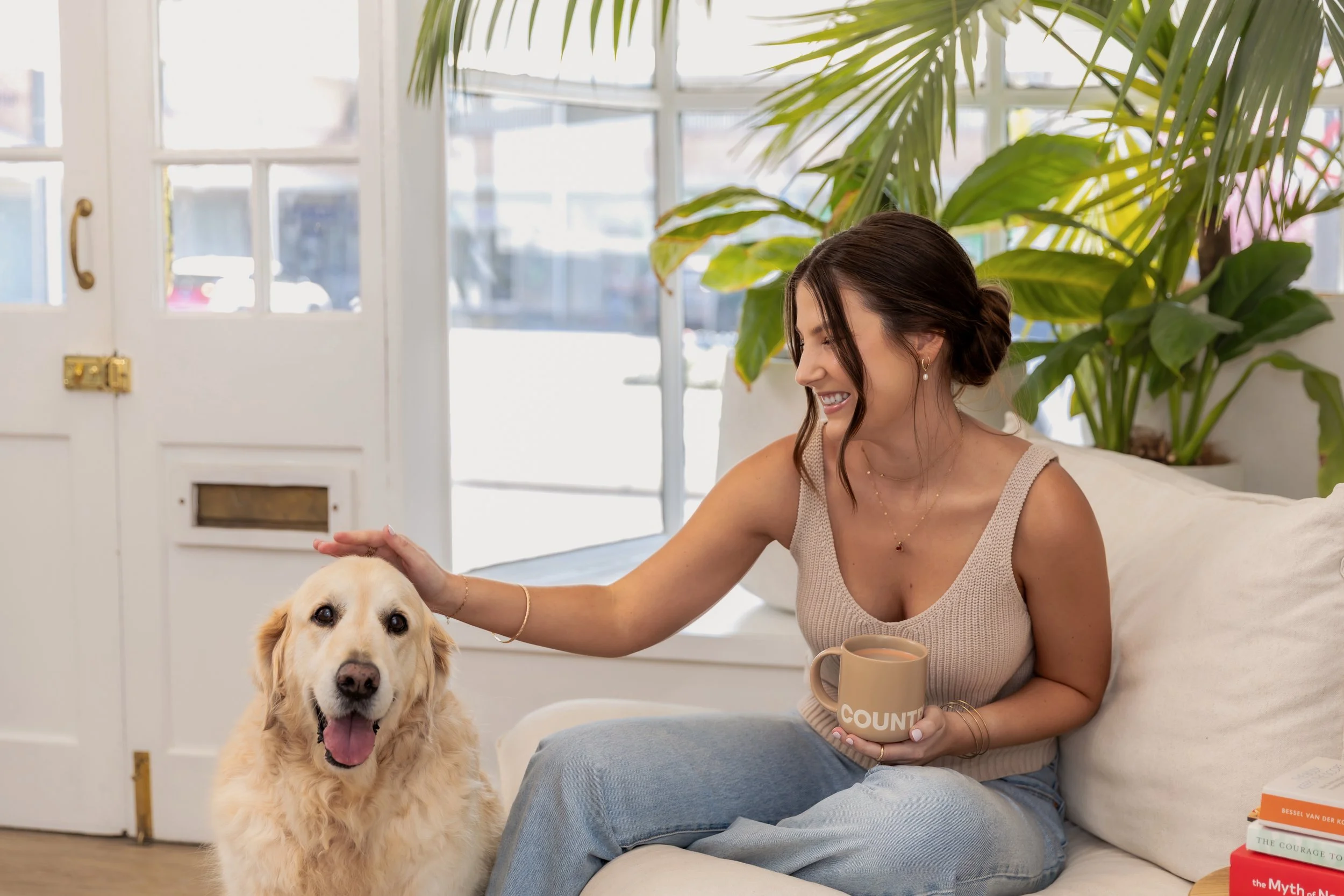 A woman sitting on a white couch, smiling and petting a golden retriever dog. She is holding a beige mug and has a large green plant behind her. The room is bright with white walls and windows.