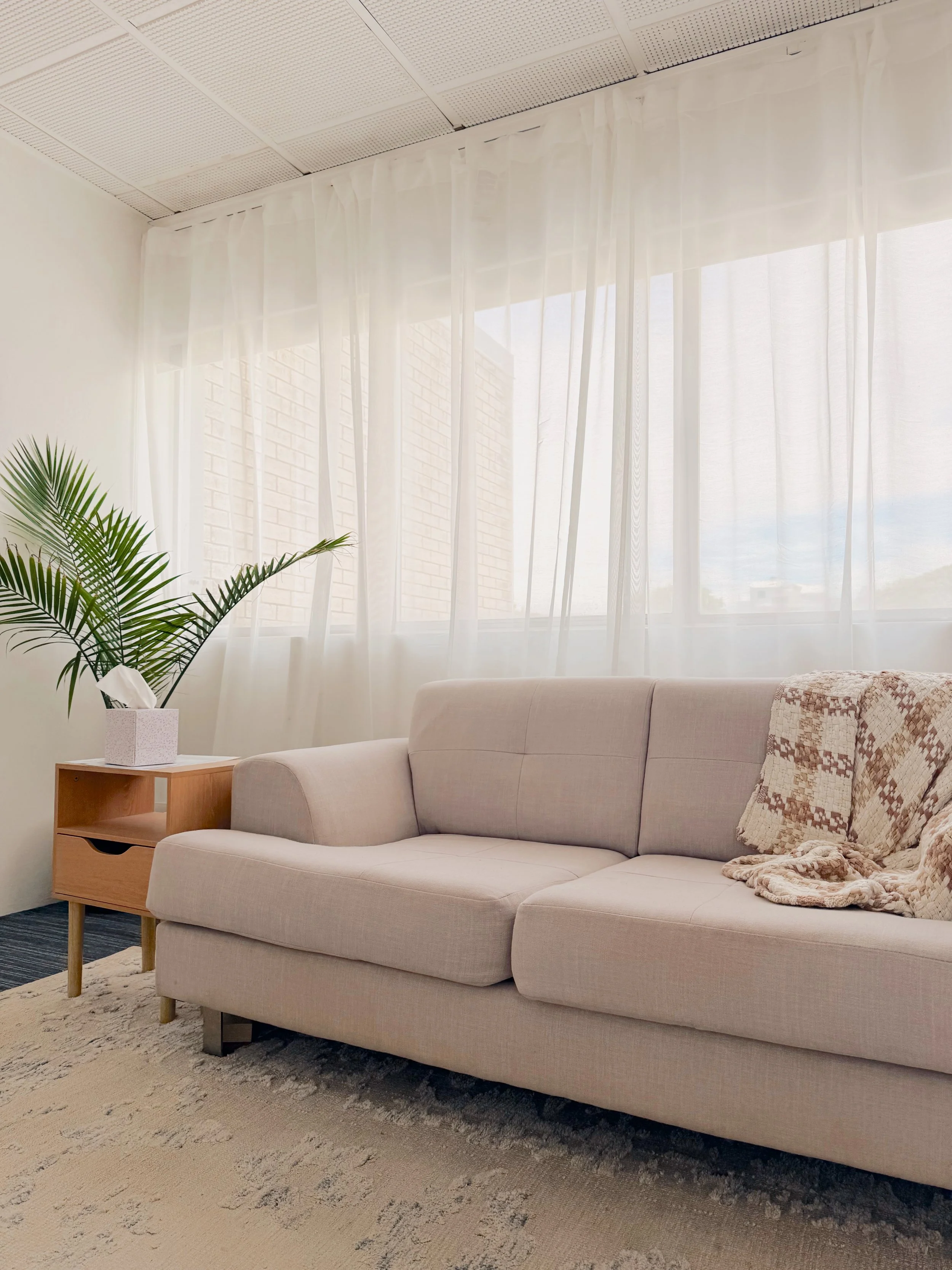 A cozy living room with a beige sofa, a small wooden side table with a potted plant and tissue box, sheer white curtains over a window letting in natural light, and a patterned throw blanket on the sofa. Carpet with a textured design on the floor.