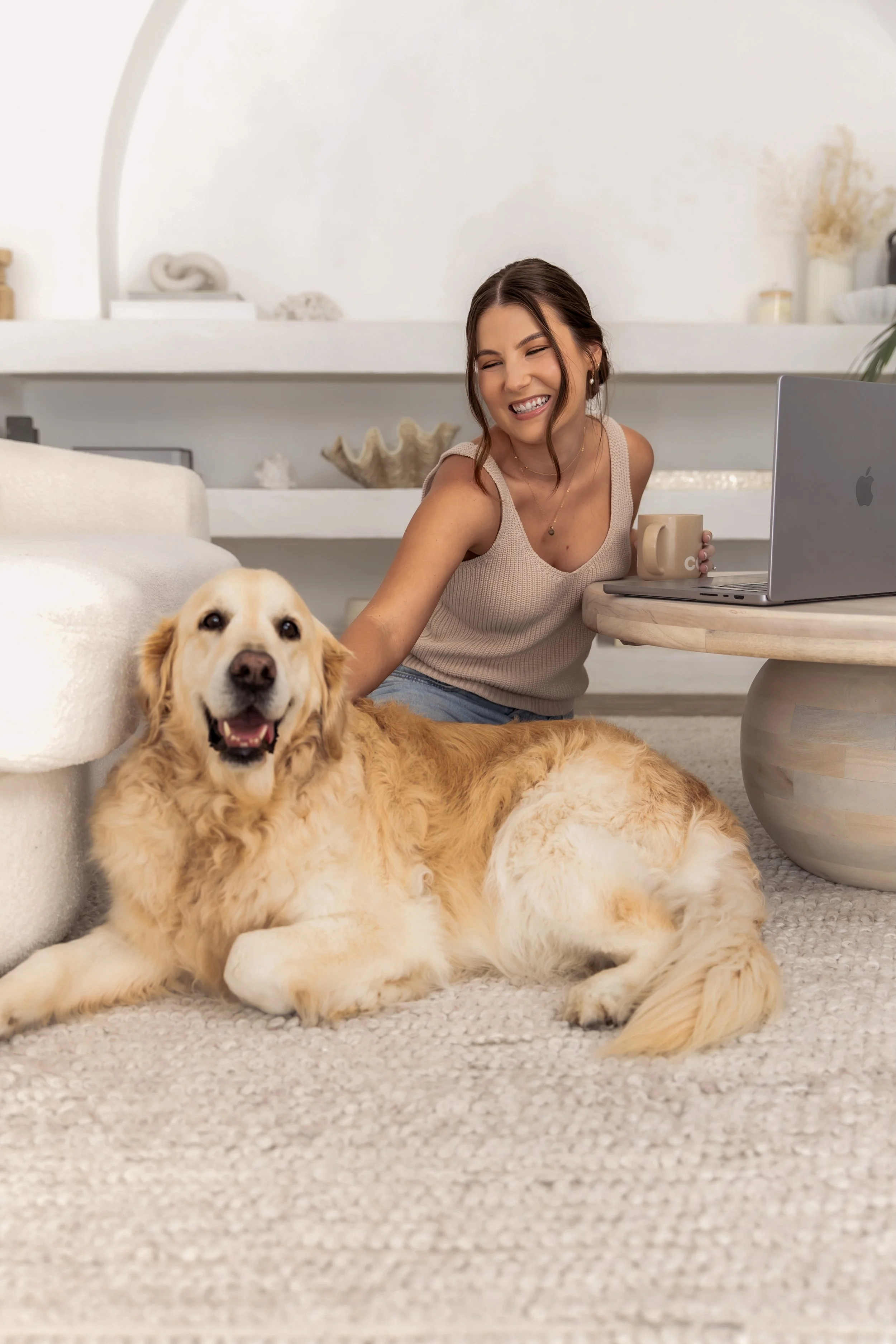 A woman sitting on a light-colored carpet with a golden retriever laying beside her. She is smiling and holding a beige mug, with a laptop on a nearby round wooden table. The background shows a white wall with decorative items.