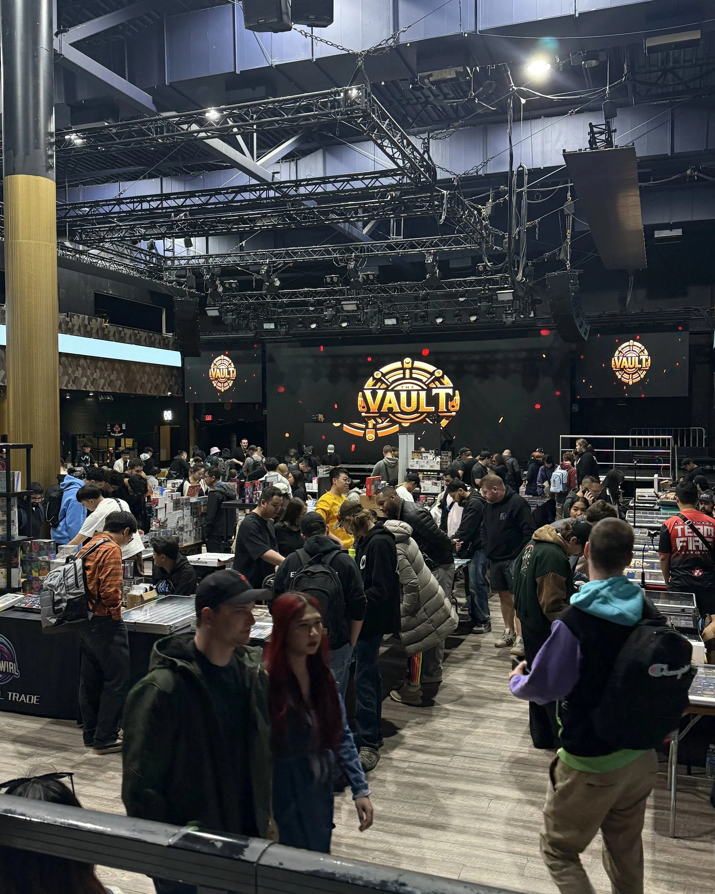 Crowd of people at the gaming event 'Vault', browsing tabletop games and merchandise in a large indoor space with a stage and screens.
