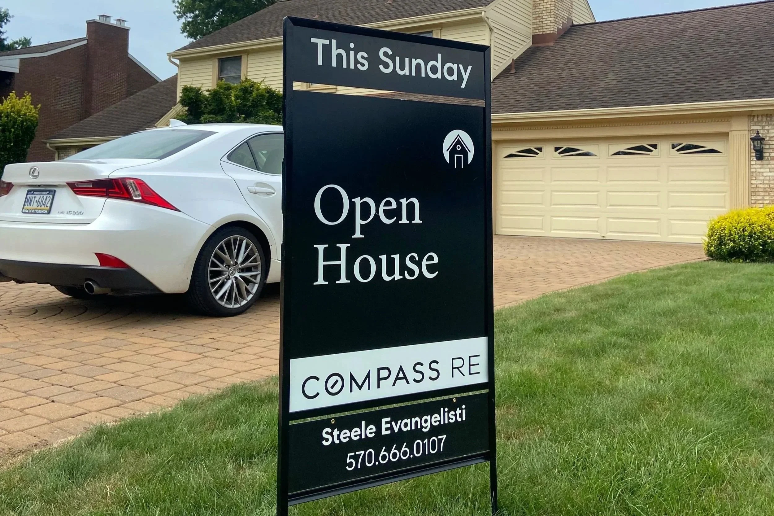 Sign outside a house advertising an open house event this Sunday, hosted by Steele Evangelisti of Compass RE, with a phone number 570.666.0107. The house features a driveway with a white Lexus sedan parked on it, a two-car garage, and the exterior is beige with brick accents. There is a well-maintained lawn and some shrubbery.