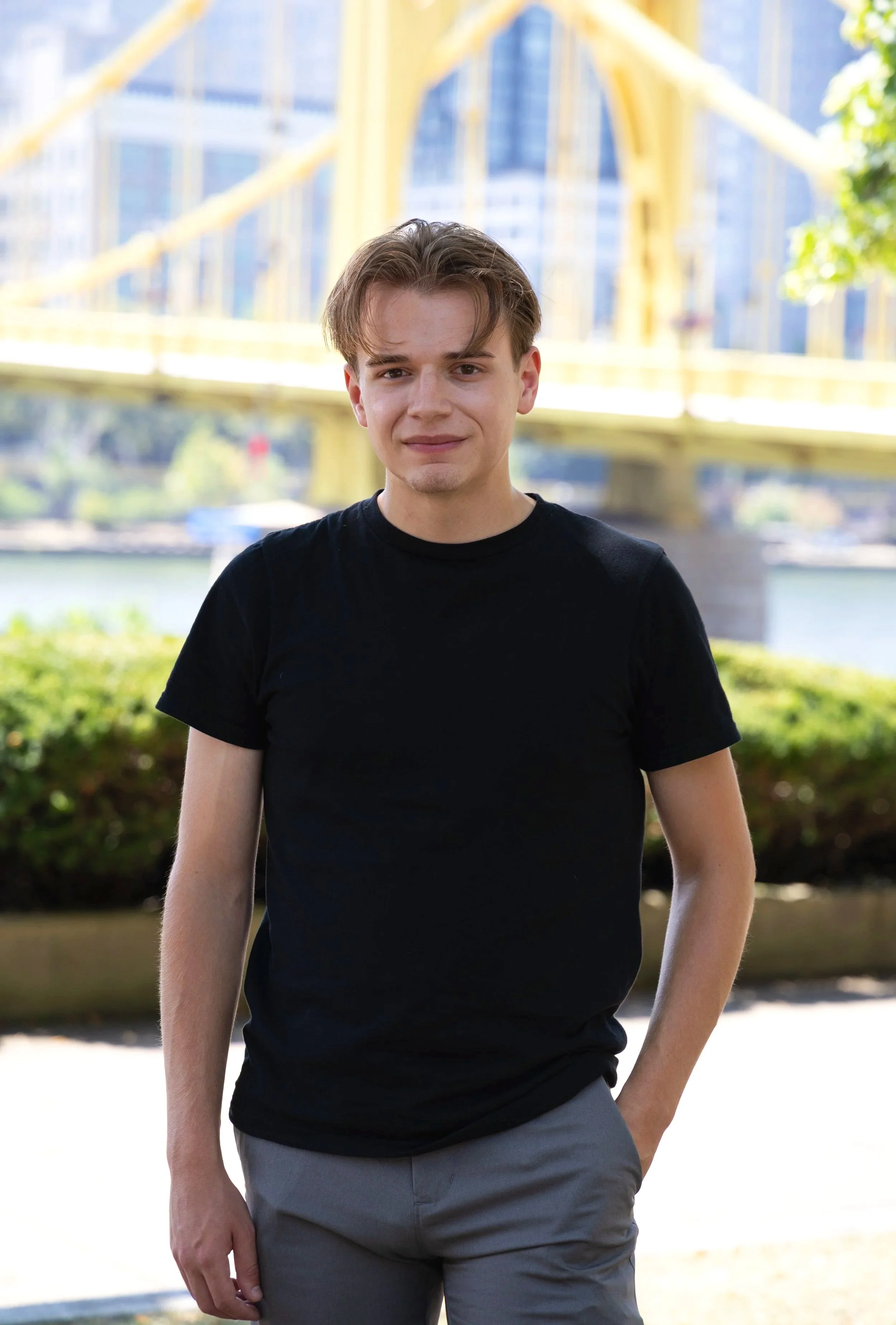 A young man in a black T-shirt and gray pants standing outdoors with a city bridge and river in the background.