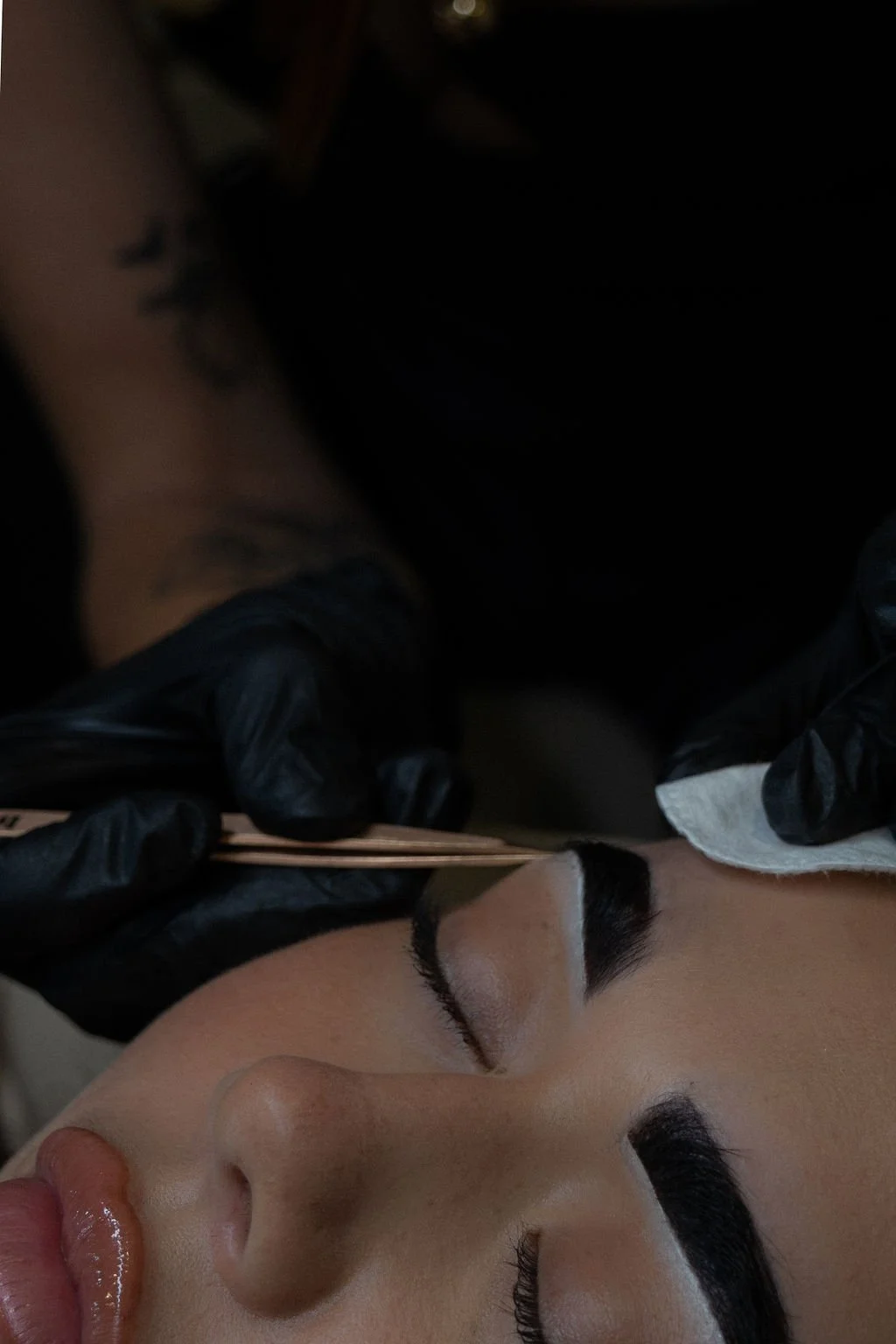 A person is receiving a beauty treatment or tattoo on their eyebrow, with another individual wearing black gloves applying a small tool or brush to the eyebrow area.