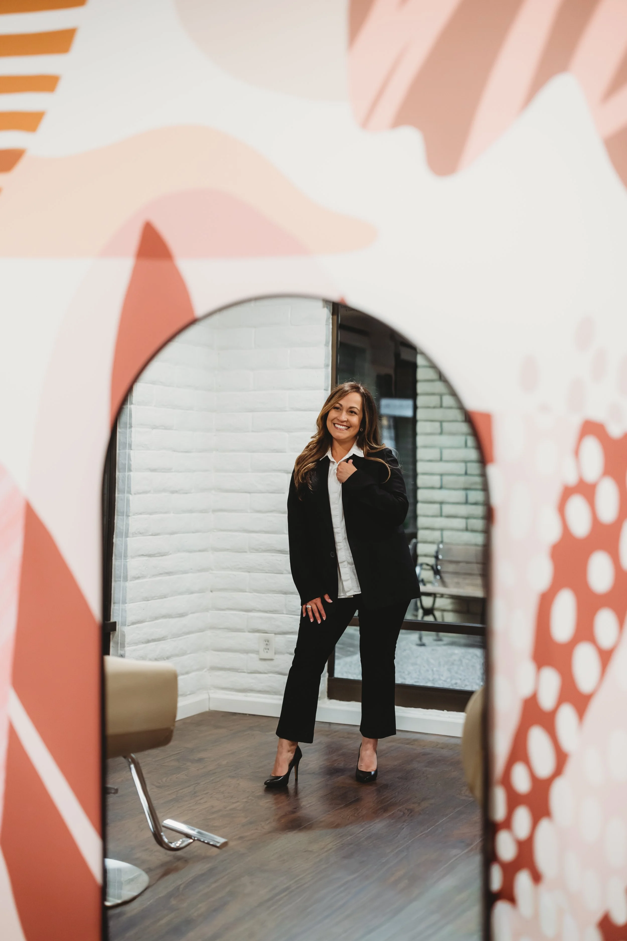 A woman standing inside a room, smiling, wearing a black suit and high heels, seen through a decorative mirror with a colorful abstract frame.