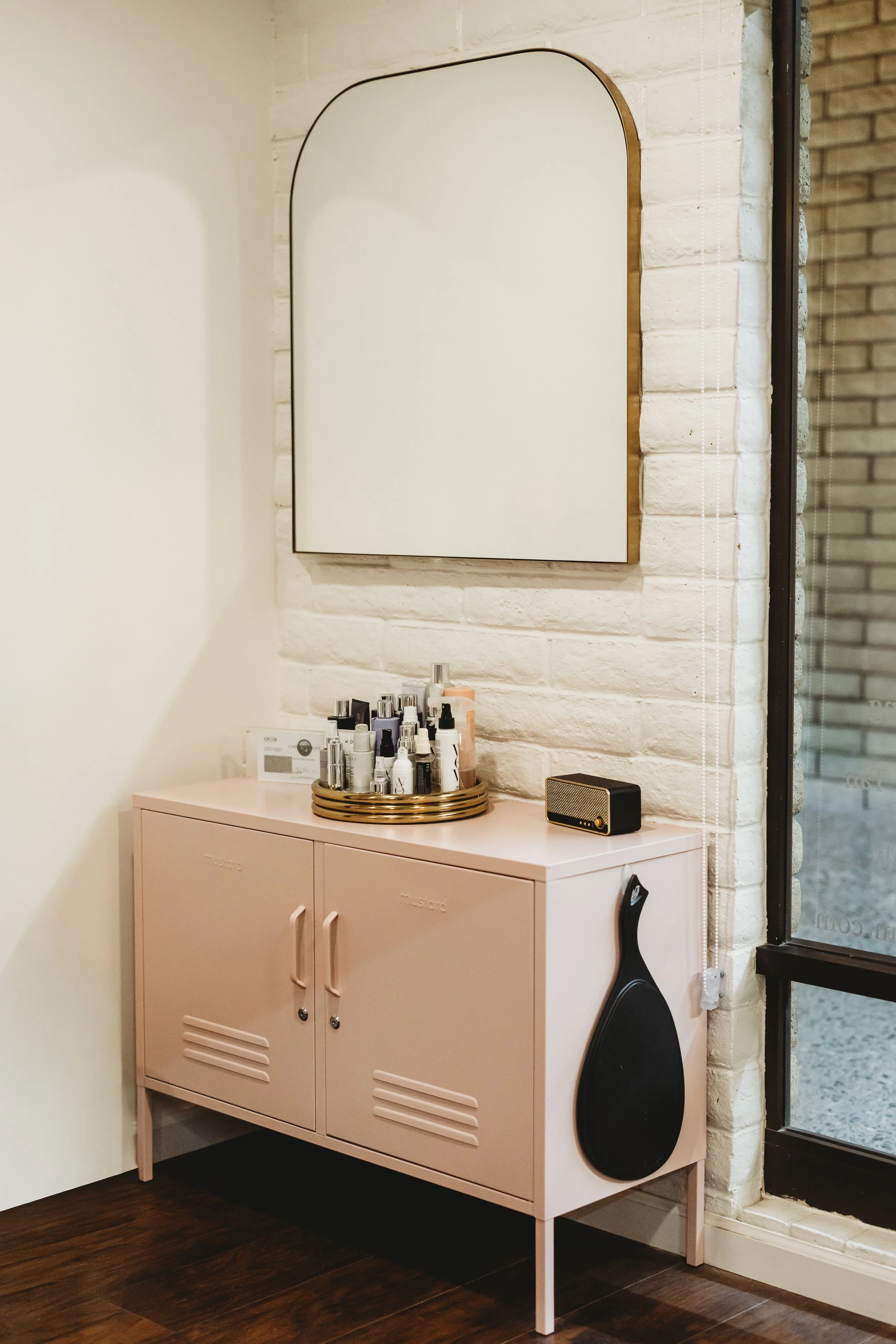 Pink cabinet with bottles of skincare and a black paddle-shaped paddle hanging on its side, next to a mirror on a white brick wall.