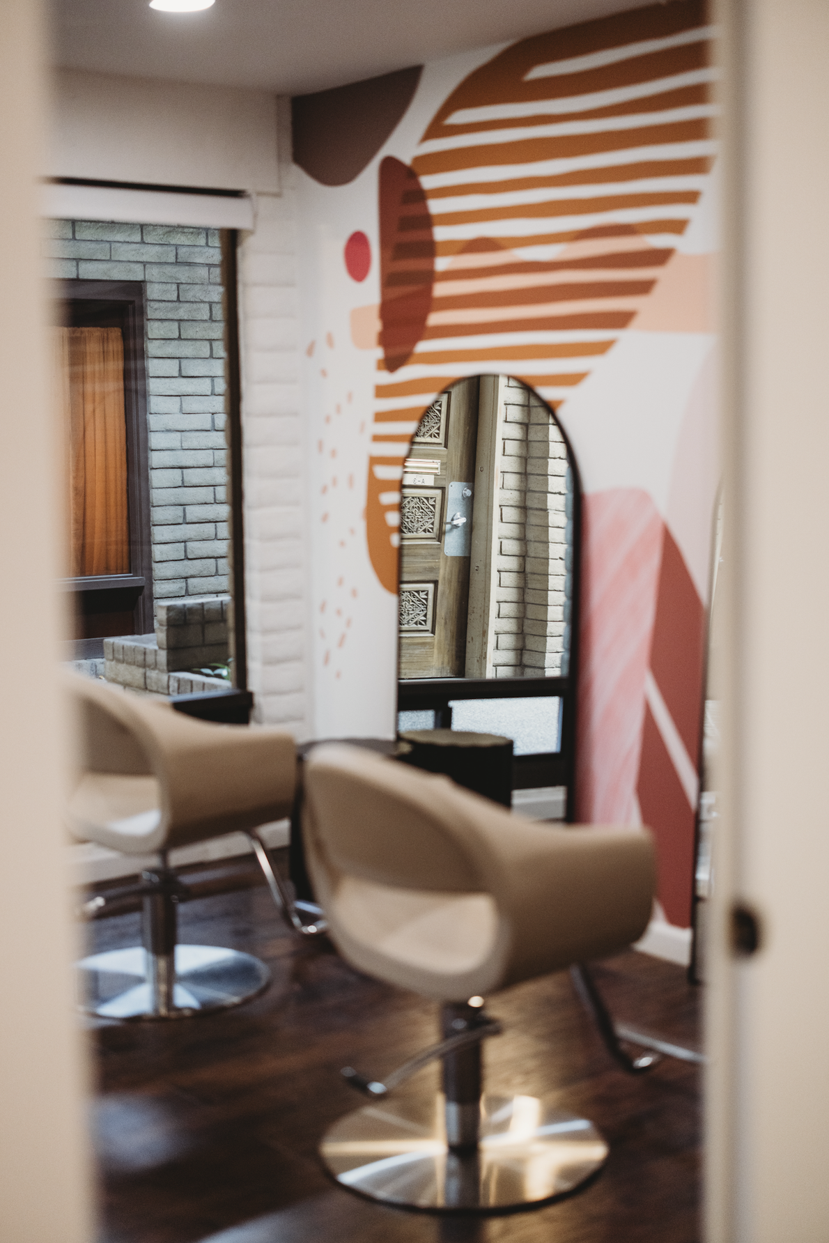 A modern salon with two beige styling chairs facing a rounded mirror on a colorful abstract wall painting, wooden flooring, brick wall, and a window with blinds.