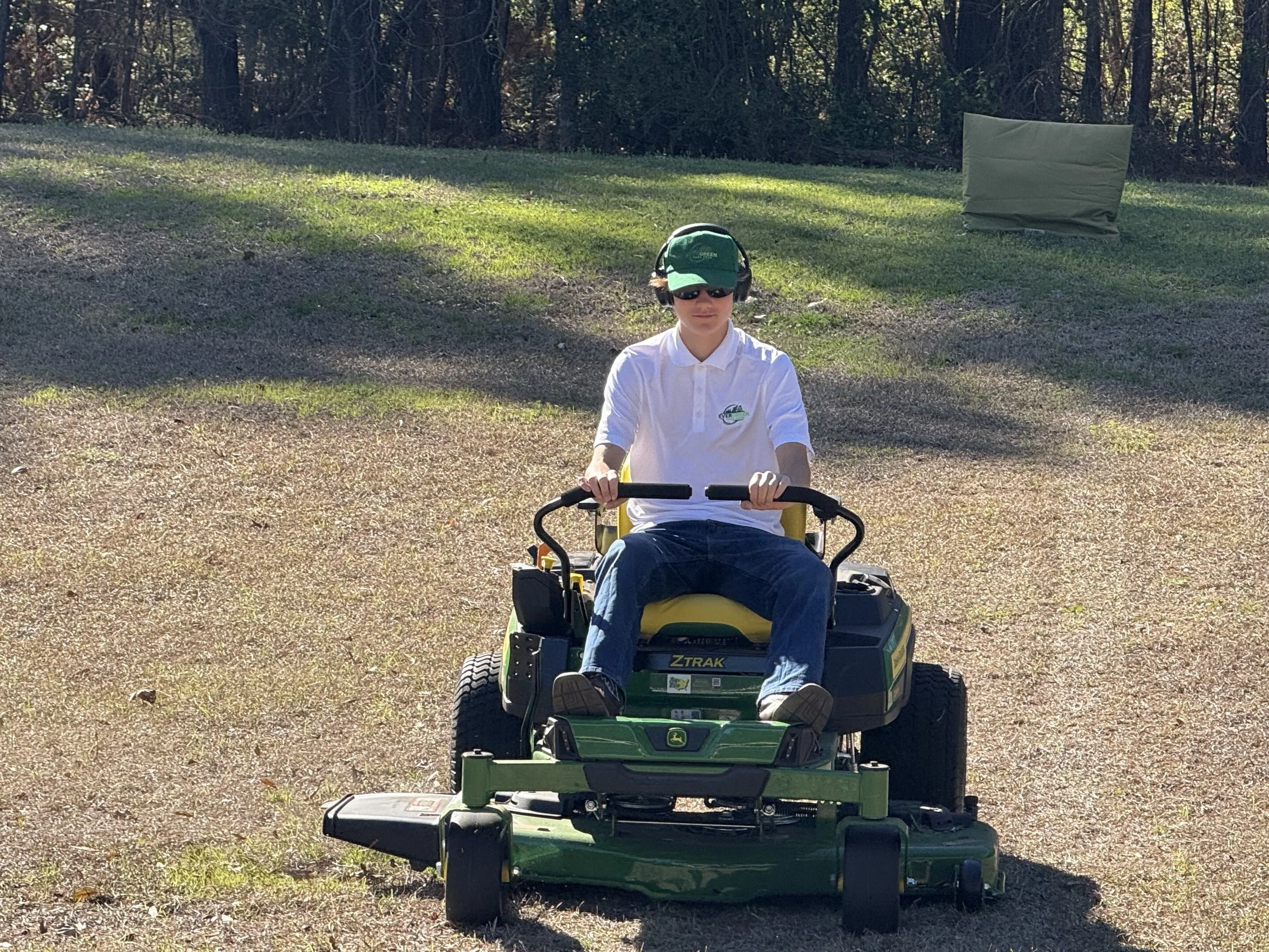 A young person wearing a white polo shirt, green cap, sunglasses, and headphones rides a John Deere Ztrak riding lawn mower on a gravel area with grass and trees in the background.