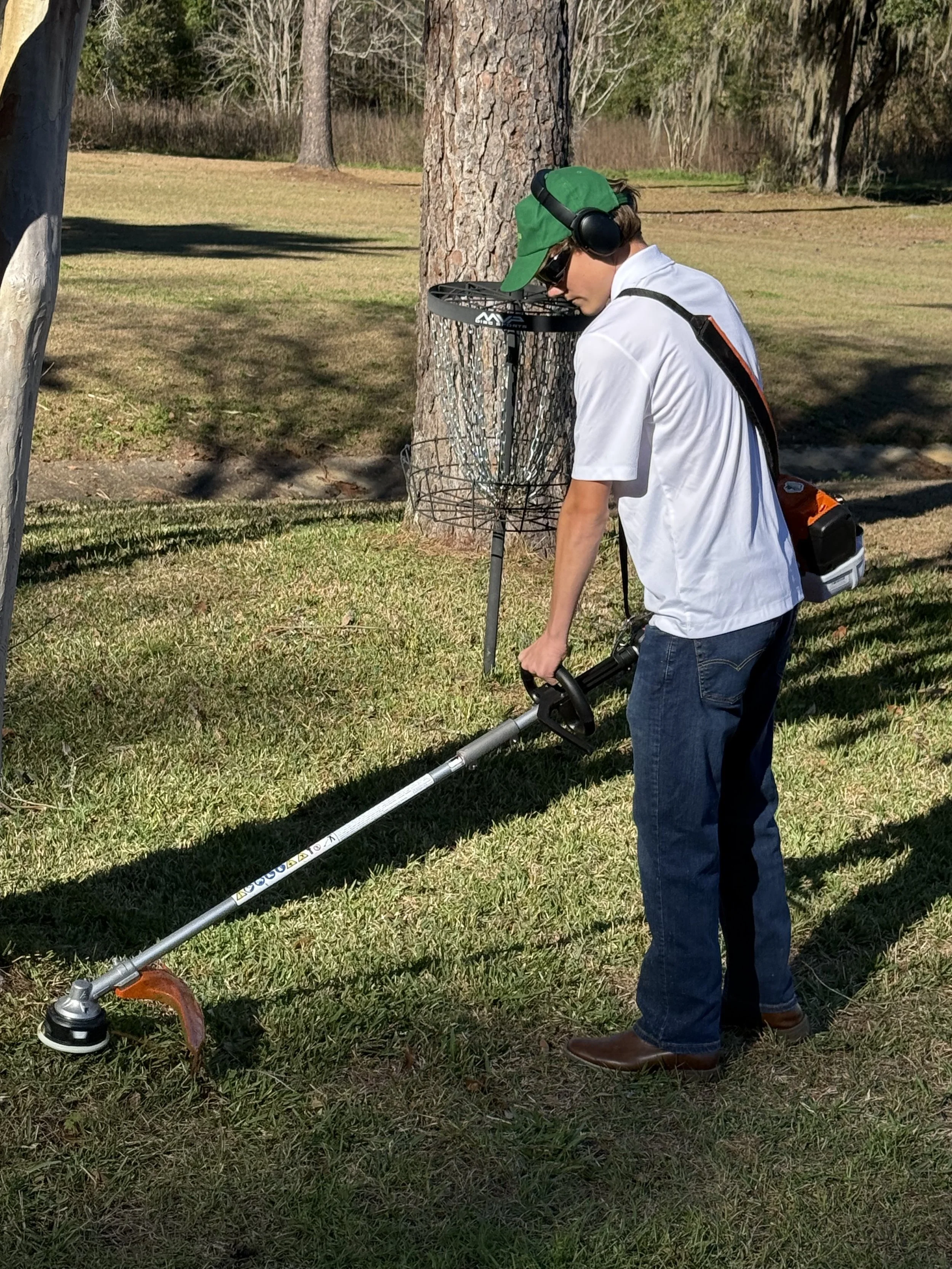 A young man wearing a green cap, sunglasses, headphones, a white shirt, and jeans is using a metal detector on a grassy area near a tree. There is a disc golf basket in the background.