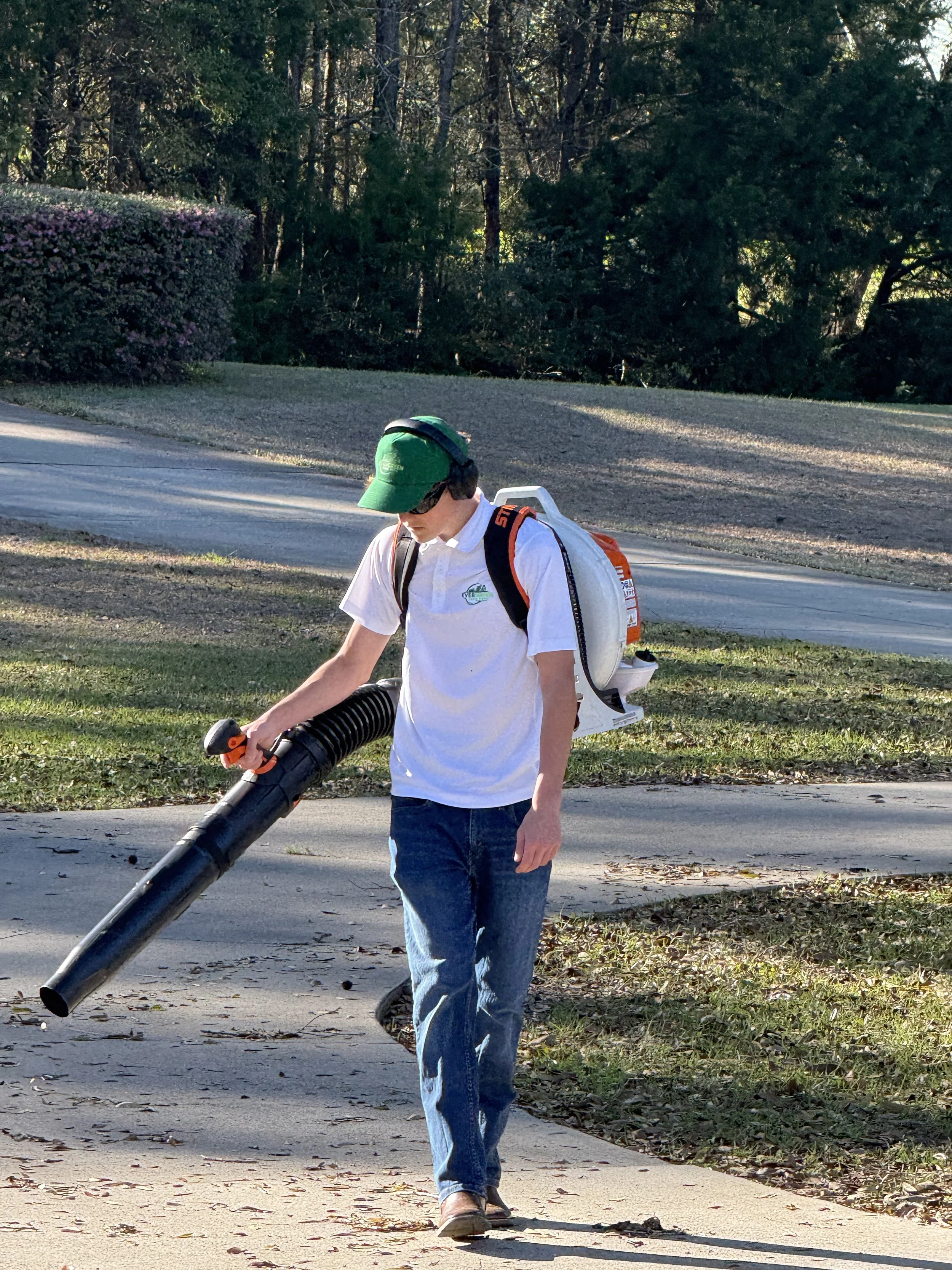 Young man wearing a green cap, sunglasses, headphones, white polo shirt, and jeans, holding a leaf blower while walking on sidewalk in a suburban neighborhood with trees and grass in the background.