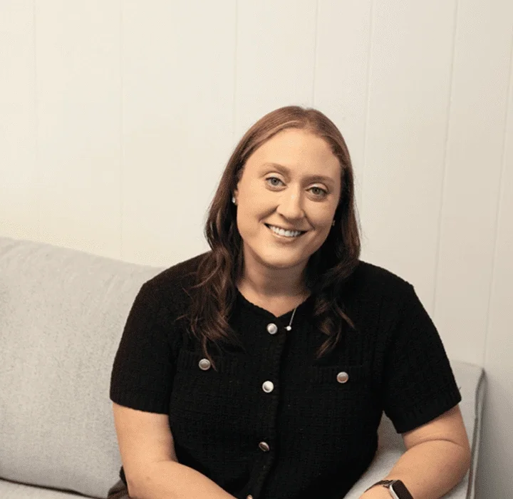 A smiling woman with long brown hair, wearing a black short-sleeved top with decorative buttons, sitting on a light-colored sofa against a white wall.