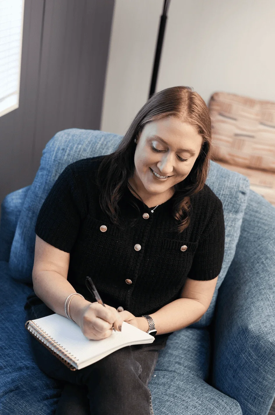 A woman sitting on a blue sofa, smiling, and writing in a spiral-bound notebook with a pen. She has long, dark hair, is wearing a black top with buttons, earrings, a watch, and bracelets.