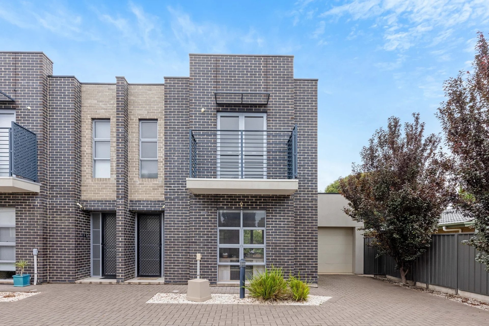 Modern two-story brick apartment building with a small balcony, large window, and front entrance, surrounded by paved driveway, trees, and a blue sky.