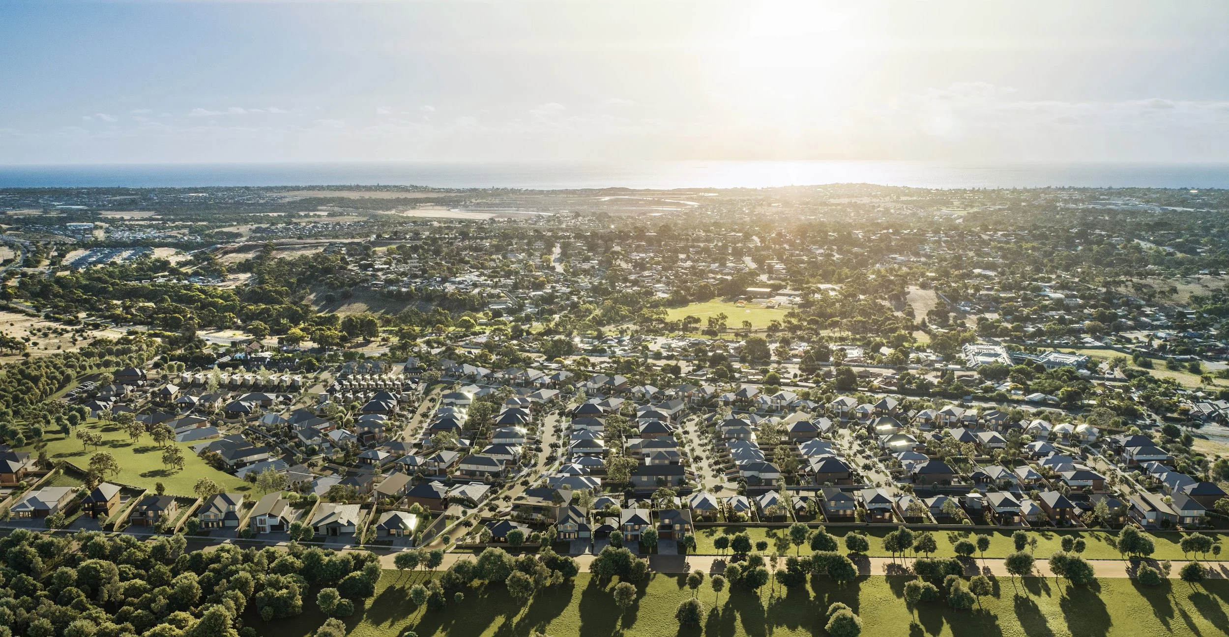 Aerial view of a suburban neighborhood during sunset with houses, trees, and green spaces, overlooking a distant city and coastline.
